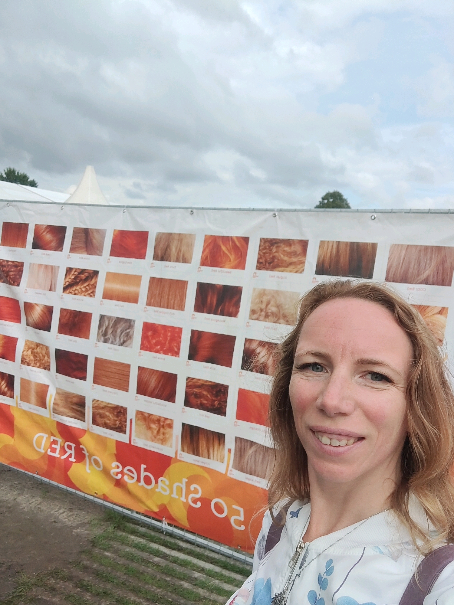 A white banner with multiple swatches of red hair colors is displayed outdoors, with a woman taking a selfie in front of it. The banner, which reads "50 Shades of RED," is partially obscured by the woman. The background shows a cloudy sky and green grass. The location is identified as the city of Tilburg, Netherlands.