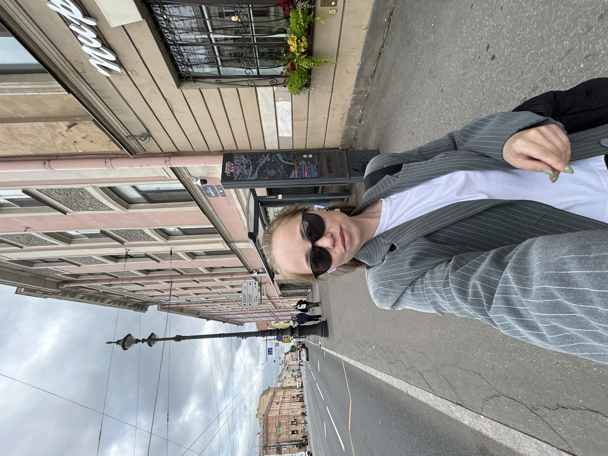 A young woman with light brown hair, wearing sunglasses and a grey pinstripe blazer over a white t-shirt, takes a selfie on a sidewalk in Saint Petersburg, Russia. She has on white wireless earbuds and her fingernails are painted a light green color. In the background, a street with traffic and pedestrian walkways stretches into the distance. Buildings with ornate architecture line the street, one of which has a sign with white cursive lettering that appears to read "Map". A lamppost stands on the sidewalk, and overhead wires crisscross the cloudy sky. The woman is positioned in the foreground, with the street and buildings occupying the background.