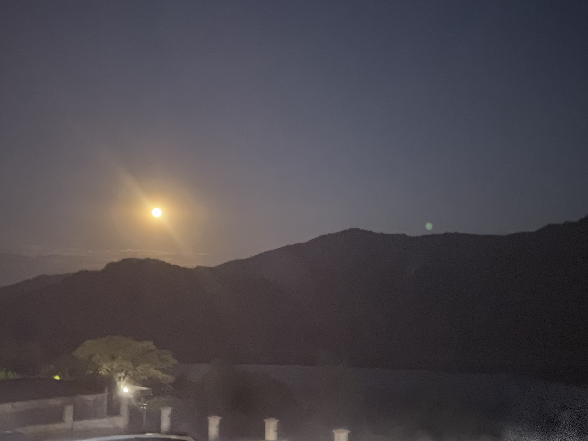 The image depicts a nighttime landscape under a moonlit sky, featuring a bright full moon casting a glow over a silhouette of mountains. In the foreground, there is a building with visible architectural details such as pillars and a wall. A large tree with illuminated foliage stands near the building, with a streetlamp nearby casting a warm light. The overall scene suggests a rural or semi-rural setting. There are no people visible in the image. The location is identified as Asparuhovo, Bulgaria.