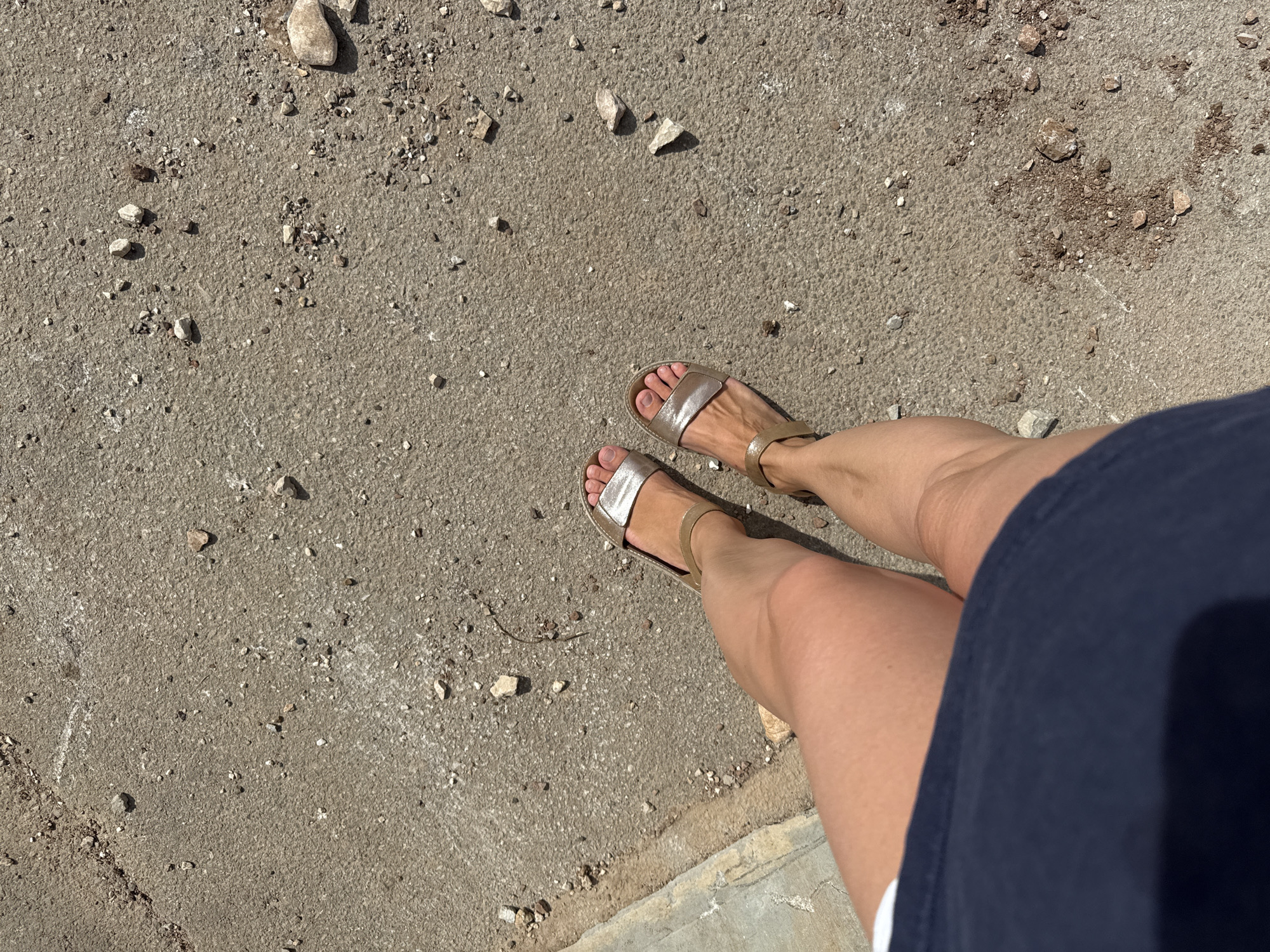 The image depicts a person standing on a paved surface, likely in an outdoor location. The surface appears to be a sidewalk or path made of concrete or asphalt, with scattered small rocks and debris. The person is wearing gold sandals. The lower part of a blue garment is visible, indicating the person is wearing a dress or shorts.
It is difficult to determine the precise location within Budva, Montenegro, from this image alone. However, the paved surface and presence of rocks suggest an area near a beach, park, or older part of the town, where such materials might be found. Without more contextual information, such as landmarks or street signs, pinpointing the specific location is not possible.
