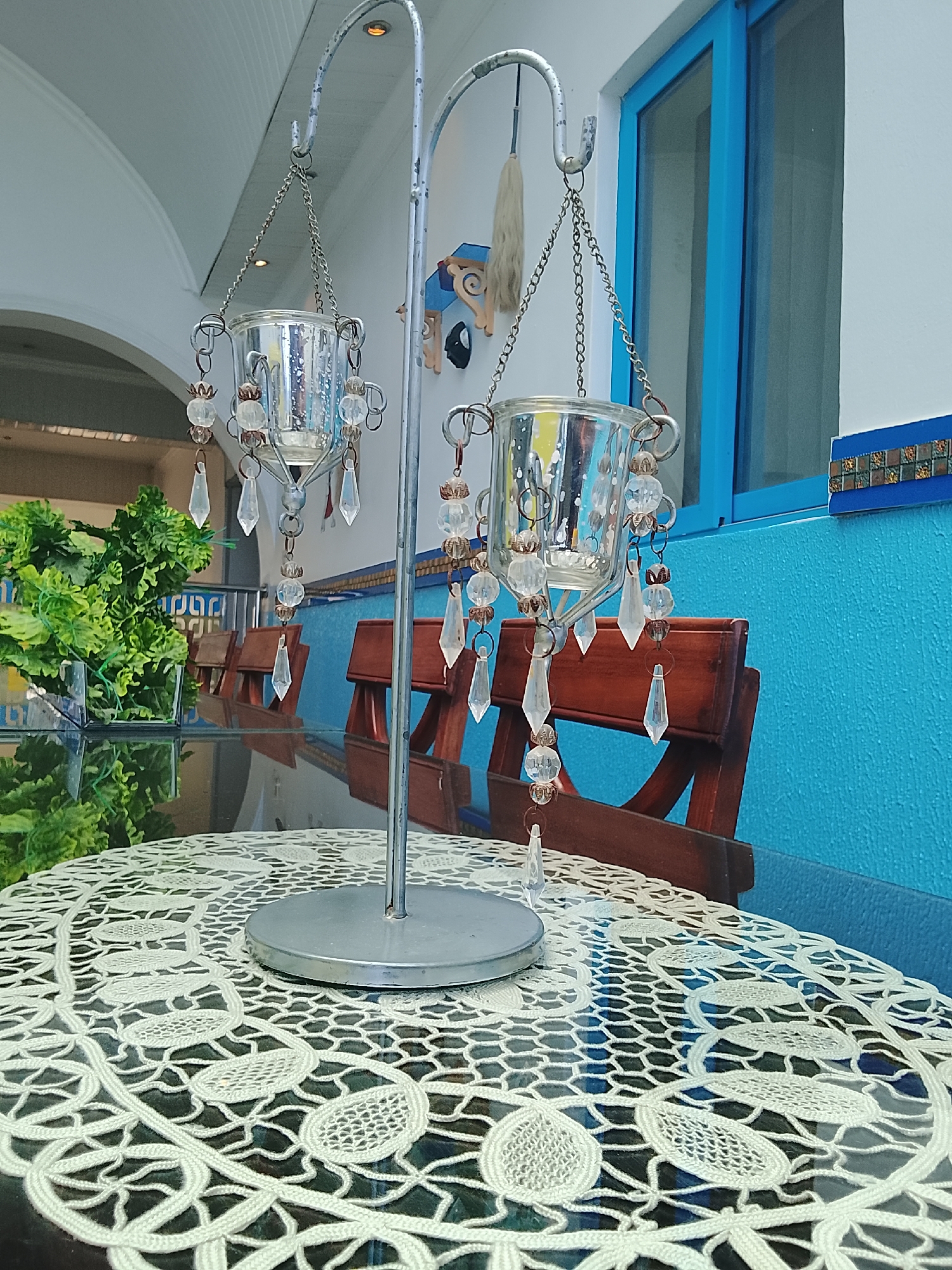 Here is a detailed description of the image:
**Scene:** The photograph captures a scene of a decorated outdoor or semi-outdoor setting. At the center is a metal stand with two hanging candle holders. Each candle holder is made of glass and adorned with decorative crystal beads and pendants.
**Objects:**
- The table underneath has a glass top covered by a lacy, crocheted tablecloth.
- Behind the table are wooden chairs.
- In the background, there is a blue wall with a window. A decorative wall hanging is seen behind the candle holders.
**Environment:**
- The scene appears to be a patio or veranda, given the presence of outdoor furniture and a view of greenery in the background.
- The location seems to be in an area with a distinct architectural style, as suggested by the painted blue walls and decorative elements.
**In summary:** The image depicts a decorated table setting on a patio or veranda, featuring a metal candle stand with crystal-embellished glass candle holders. The scene is set against a blue wall and outdoor greenery, suggesting a location with a distinct architectural or regional character.