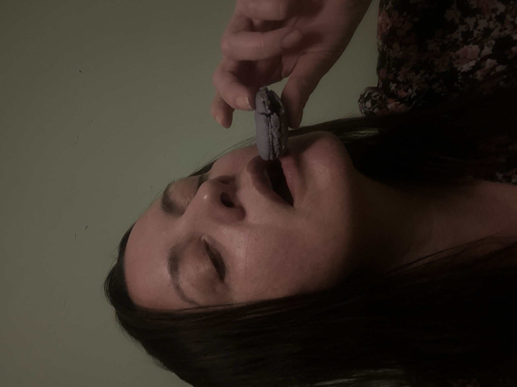 A medium shot depicts a person with closed eyes, lying on their back with dark hair framing their face. Another person’s hand is shown holding a partially bitten, cracked purple macaroon directly over the first person's slightly open mouth. The setting appears to be indoors, against a plain green wall. There is no information to identify the location in Riga, Latvia.
