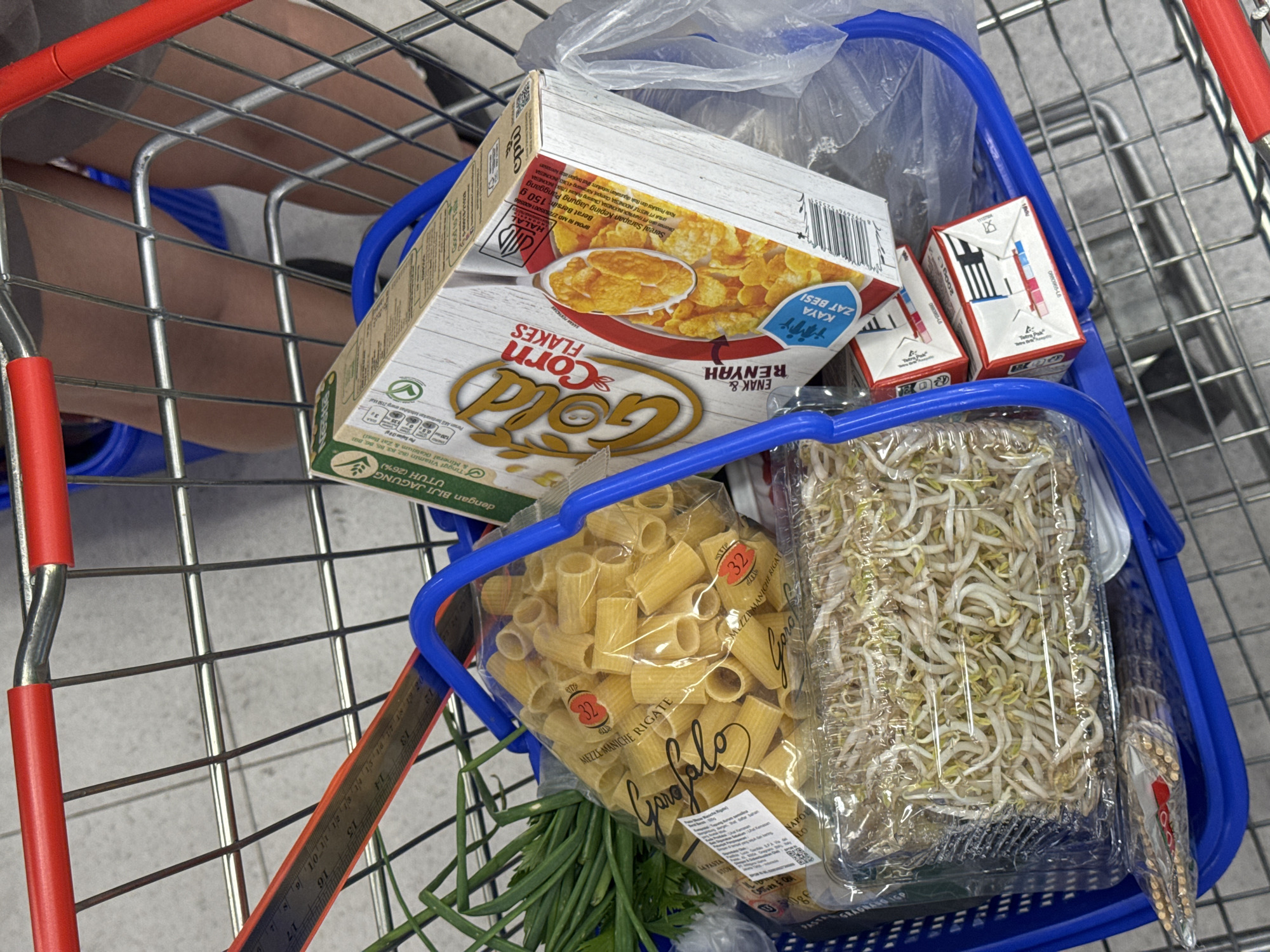 A shopping cart contains a blue basket filled with grocery items. The items include a box of Gold Corn Flakes cereal, two small Tetra Pak cartons, a package of Garofalo Mezze Maniche Rigate pasta, a package of bean sprouts, and green onions. A ruler is visible near the bottom left corner of the basket. The shopping cart's handle is red. A person's legs and feet are visible in the upper left portion of the image. The location is unknown but the boxes of cereal mention Indonesia. The text "PT NESTLE INDONESIA, Campo Karawang 413 INDONESIA" can be seen on the box. There is no reference to Kuta in the texts.