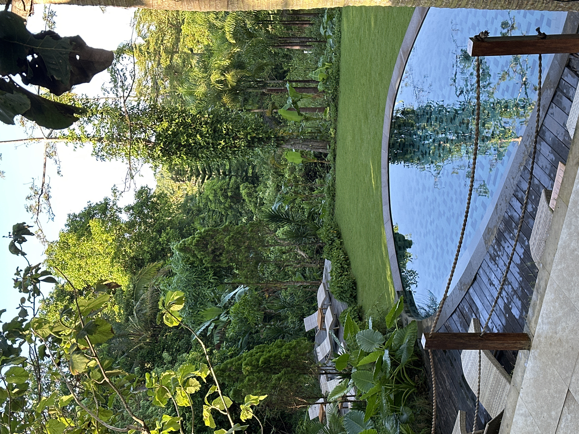 The image captures a serene scene in Nusa Penida, Indonesia. A kidney-shaped pool with light blue tiling is bordered by a wooden deck and lush green lawn. Beyond the lawn, a dense tropical forest rises, featuring a variety of trees and vegetation. Lounge chairs are visible on the deck near the pool, with ropes hanging down from a wooden beam above. The photograph appears to have been taken during the day, with natural light illuminating the environment and reflecting off the water's surface.
