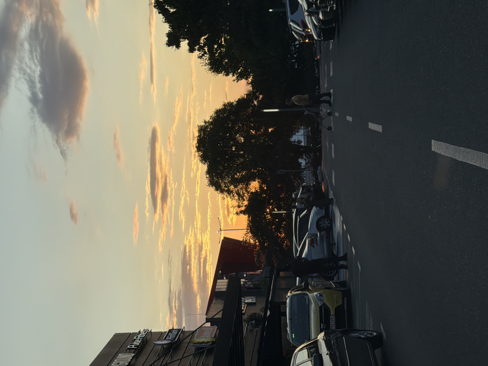 The image depicts a sunset scene in the parking lot of a shopping center in Burgas, Bulgaria. The sky is a gradient of orange, yellow, and pale blue, with scattered clouds. In the foreground, the asphalt parking lot is marked with white lines. Several cars are parked, including a dark sedan on the left, a yellow Dacia Logan in the center, and several other vehicles further back.
People are present in the parking lot. On the left, a person in a dark shirt and pants stands near the yellow car. Further to the right, a person in a yellow shirt and dark pants is pushing a shopping cart. In the background, more people are visible near the parked cars.
A building with signage is visible on the left, appearing to be a commercial establishment with "GROUND Pulse Fitness" and "Postbank" visible. A red angular structure is also part of the building's architecture. Trees are silhouetted against the sunset on the right side of the image. The overall atmosphere is that of a typical evening scene in a Bulgarian urban environment.
