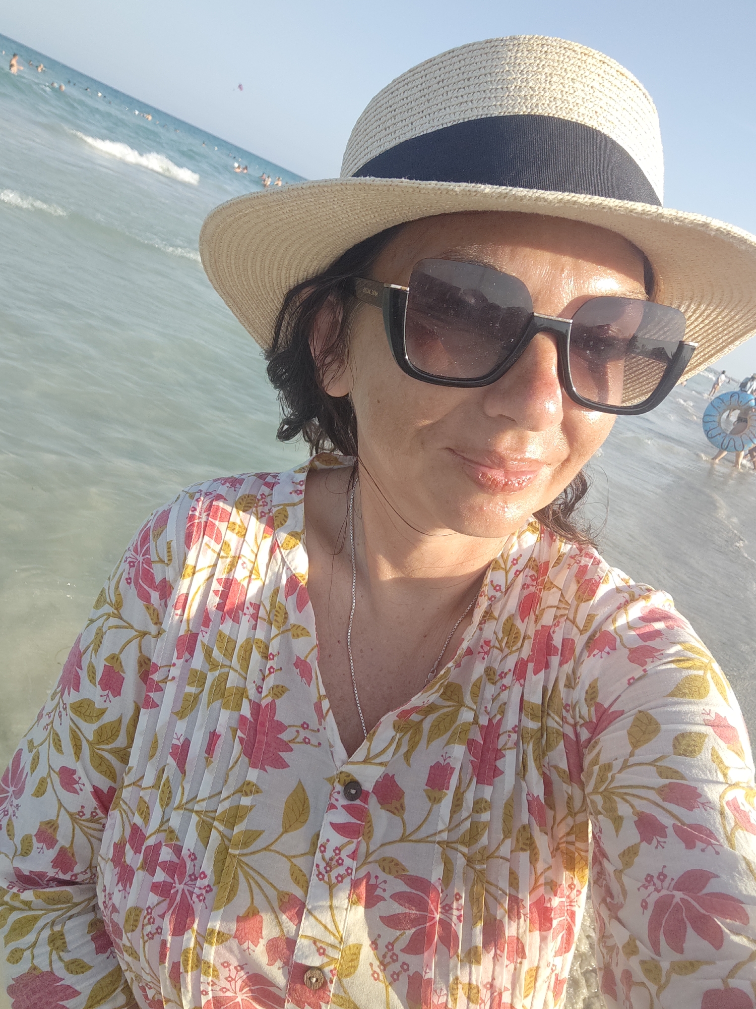 The image is a close-up selfie of a woman wearing a straw hat and sunglasses, standing in shallow water at the beach. She is wearing a white floral print blouse with pink and yellow flowers. In the background, the sea is visible with gentle waves and other beachgoers. The sky is clear and blue. The scene is likely the beach in Mahdia, Tunisia, given the presence of people enjoying the water and the clear sky.