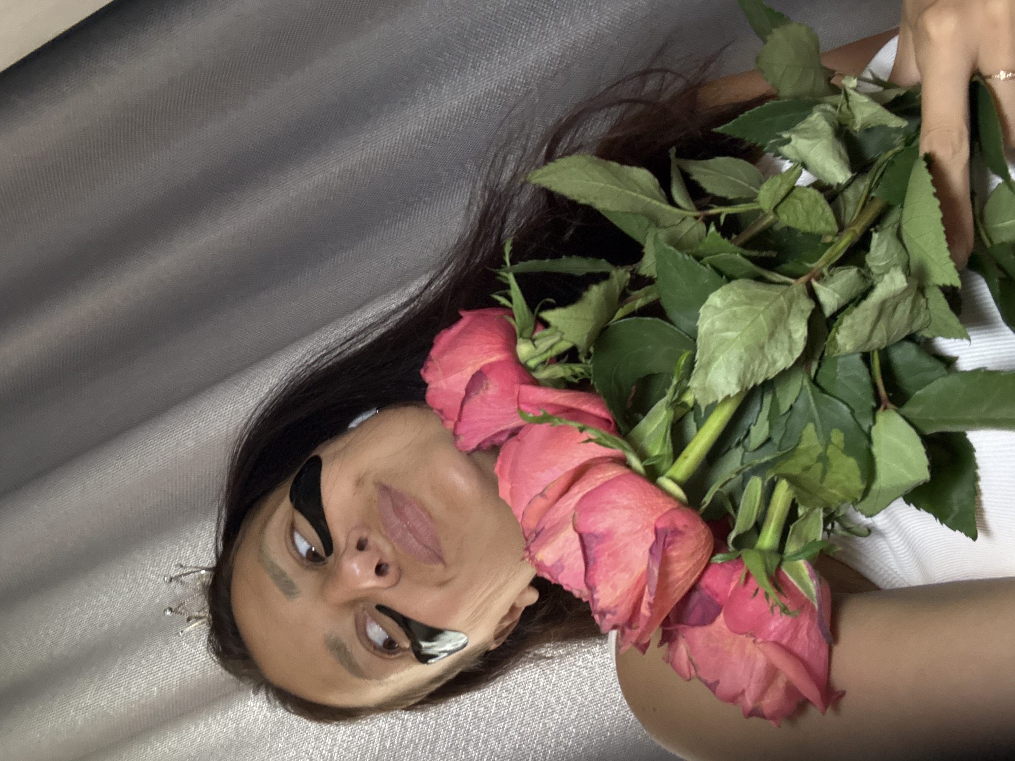 A woman with dark hair and black eyeliner posing with pink roses. She is lying on a gray textured surface and is wearing a small silver crown. Her right hand is holding a bouquet of roses. The image could be located in the city of Chaikovsky, Russia, but there is no confirmation of this location in the image itself.