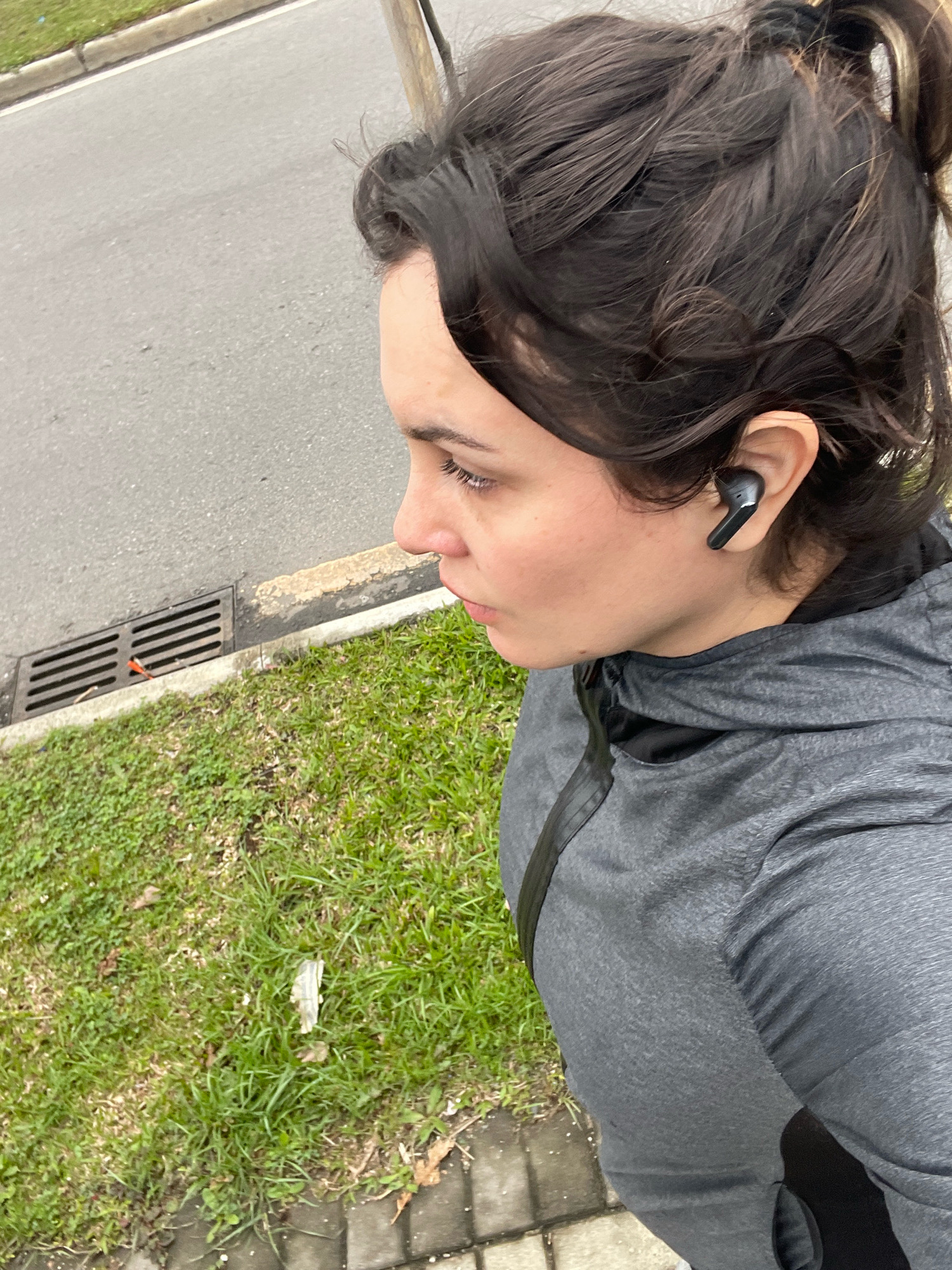 The image shows a close-up of a person's head and torso, from the shoulders up. The person is looking to their left and has dark hair pulled back in a ponytail, with a few strands loose around their face. They are wearing black wireless earbuds in their right ear and a grey hooded jacket.
The background consists of a paved road, a grassy verge, and a drainage grate. The lighting suggests it is daytime, possibly overcast. The overall scene is outdoors, on what appears to be a sidewalk or path next to a road, with grass on the side. This could be a residential street or a park area in Curitiba, Brazil, given the context. There is no visible indication of the specific location within the city itself.
