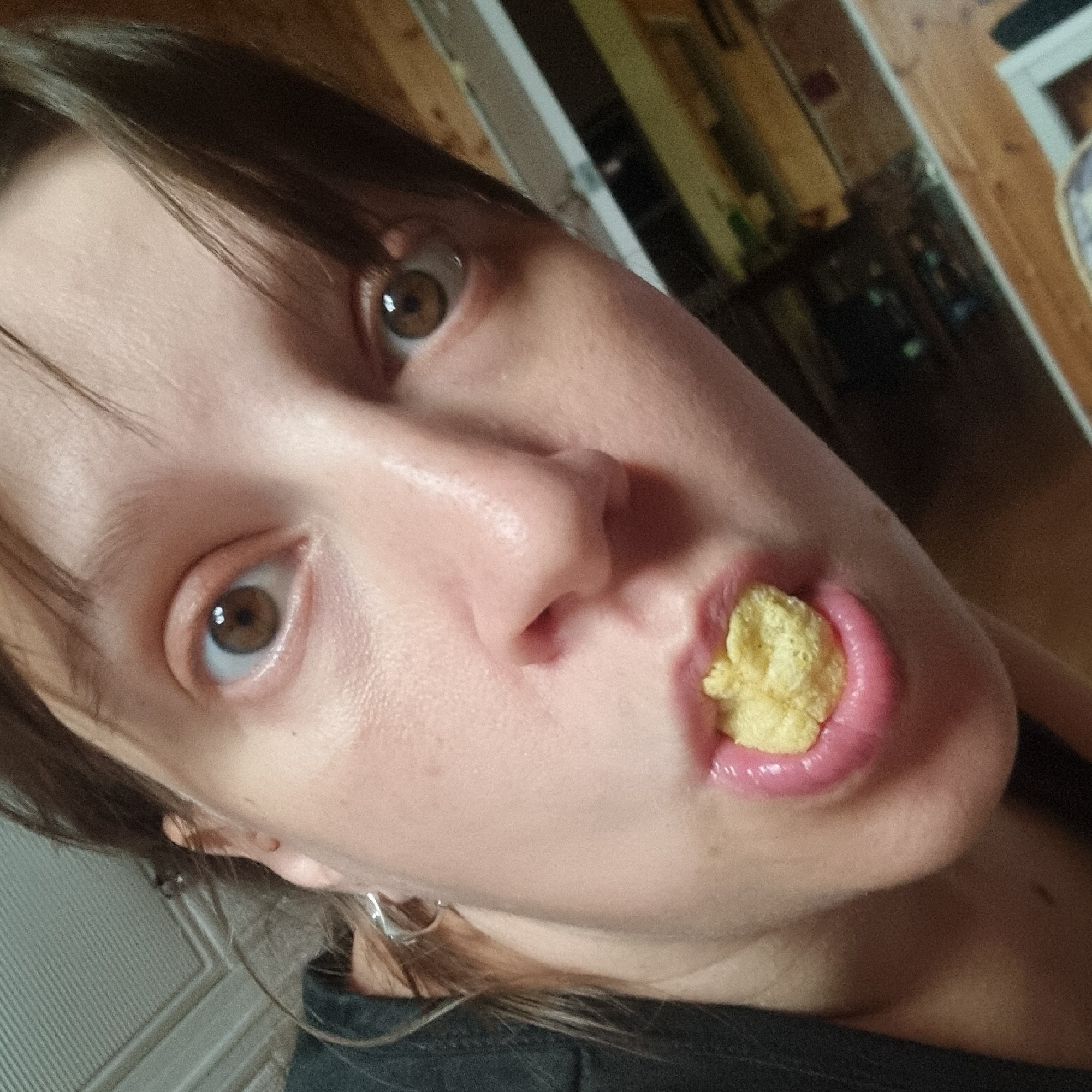 The image is a close-up, indoor, low-angle shot of a person's face, focusing on their mouth, nose, and one eye. The person has brown eyes and is holding a yellow, puffy food item in their mouth. Their lips are slightly parted and appear to have a glossy sheen. The background is blurred and shows wooden paneling and possibly shelving with indistinct objects. The image was taken in Dunnae-myeon, South Korea.