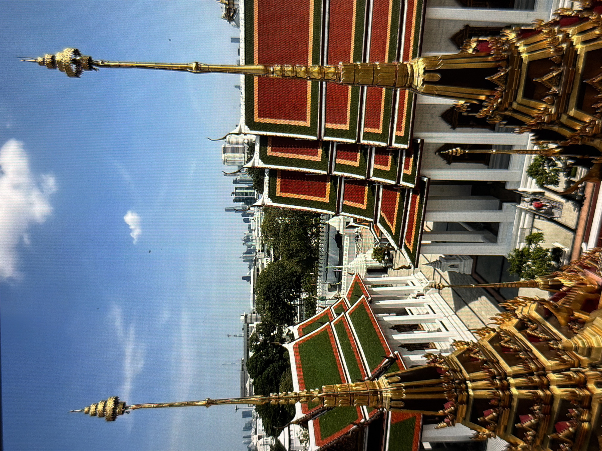 The image shows a view from a high vantage point overlooking a cityscape, likely in Bangkok, Thailand. In the foreground are ornate, gold-colored temple rooftops with red and green tiles, characteristic of Thai architecture. These roofs are elaborately decorated with tiered structures and pointed spires. Beyond the temple structures, a cityscape is visible, featuring a mix of high-rise buildings and green trees. The sky is blue with scattered clouds. There are no people visible in the image. The location is not in Đà Nẵng, Vietnam.