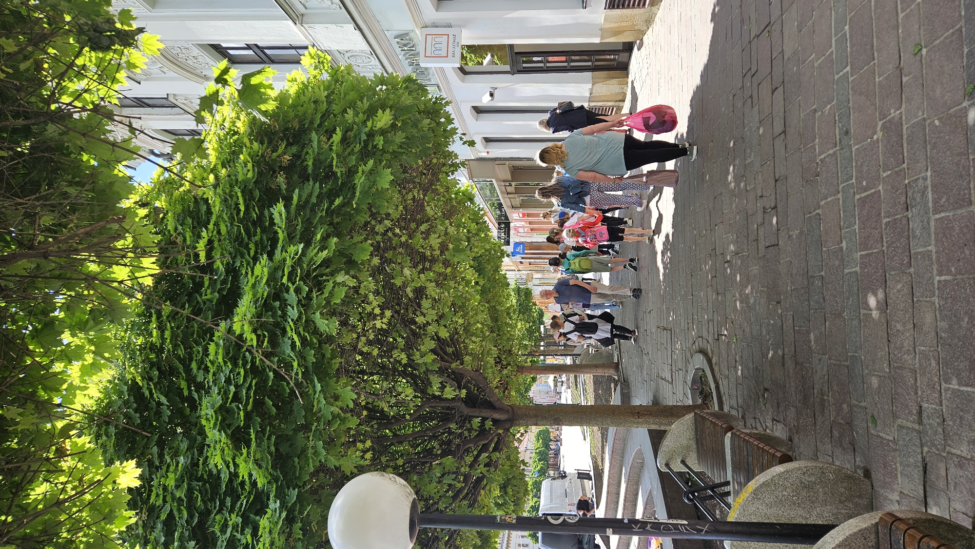 The image captures a street scene in Prešov, Slovakia. A group of people, including children and adults, are walking along a pedestrian zone paved with brick. The scene is dominated by a line of lush, green trees along the sidewalk. A building with ornate architectural details is visible. A streetlight with a spherical lamp is located on the street. Benches line the sidewalk. People are engaged in the act of walking. A bank sign is visible.