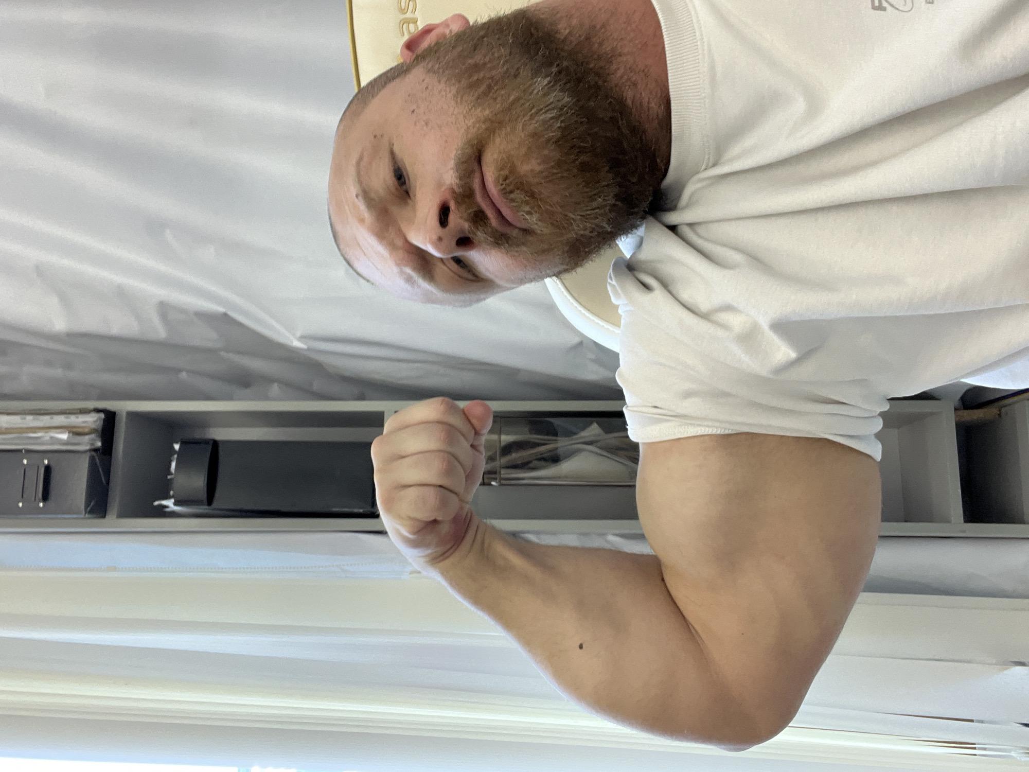 A Caucasian male with a close-cropped reddish-brown beard and receding hairline is facing the camera. He is wearing a white t-shirt. His right arm is flexed, displaying the bicep. Behind him, a shelf contains items including what appears to be a black flashlight or tube-shaped object and a box. Below the shelf, there is a white surface with faint horizontal lines, likely a window covering. There is no indication of location in Chaikovsky, Russia or any city.
