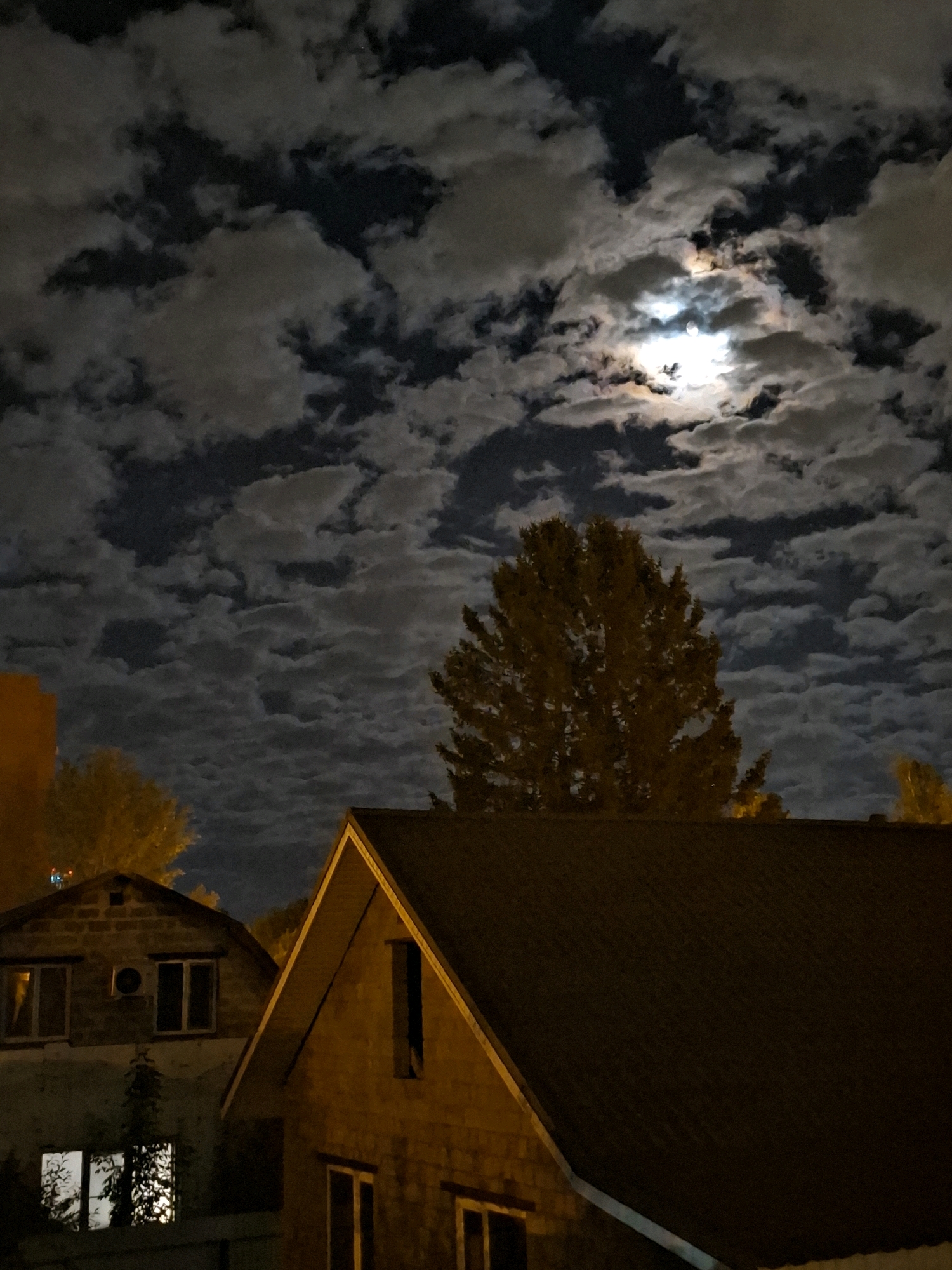 This is a nighttime, low-angle, full-frame photograph of a residential area under a cloudy sky illuminated by the moon. The scene appears to be in the city of Tyumen, Russia, based on contextual information.
In the foreground, the dark, sloped roofs of two houses dominate the lower portion of the frame. The house on the right has a prominent gable with a dark roof. The house on the left, built of brick, has multiple windows, some of which are illuminated from within, casting a warm glow onto the exterior. An air conditioning unit is visible on the brick house, as is a vine growing up its side.
Behind the houses, a large, leafy tree with yellowing foliage stands prominently in the center of the image. To its left, another tree with bright yellow leaves is partially visible, and further to the left, the upper portion of an orange building rises.
The sky above is filled with a dense layer of textured clouds, which are brightly lit by the full moon. The moon itself is visible through a break in the clouds, appearing as a luminous orb with some haloing. The interplay of light and shadow from the moon creates a dramatic effect on the clouds. The overall impression is one of a calm, cool night in a suburban or semi-rural part of the city.
