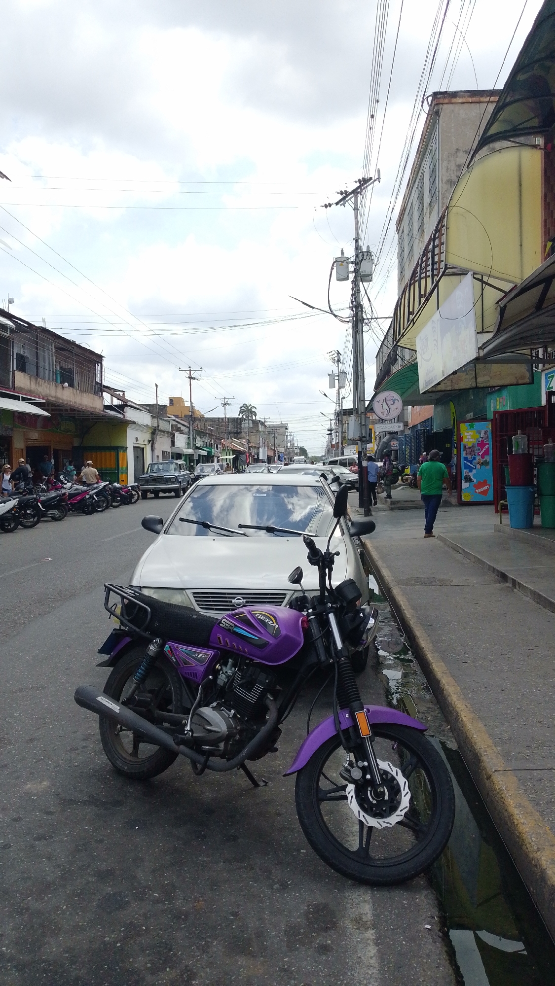 This image captures a bustling street scene in Tinaquillo, Venezuela, under an overcast sky during daytime. In the immediate foreground, a vibrant purple motorcycle is parked on the asphalt, with its front wheel resting near a yellow-painted curb and a water-filled gutter. "150" and "DER TERRAR" are visible on its side, along with its exposed engine and a front disc brake.
Behind the motorcycle, a silver Nissan sedan is parked. The street extends into the background, densely lined with parked vehicles, including numerous other motorcycles along the left-hand sidewalk. Buildings with commercial establishments on their ground floors flank both sides of the road. On the right sidewalk, a person in a green shirt and blue pants walks away from the viewer. Shop signs are visible, including "MESA.OASIN" and "BIBARES," and a colorful advertisement for "TODACASA" or "TODACASA KIDS."
Above, a complex web of utility poles and power lines crisscrosses the sky. The scene portrays the daily activity of a typical street in a Latin American town, with pedestrians, parked vehicles, and local businesses, all contributing to a sense of everyday life. No specific event is taking place; it's a snapshot of the ordinary rhythm of the town.