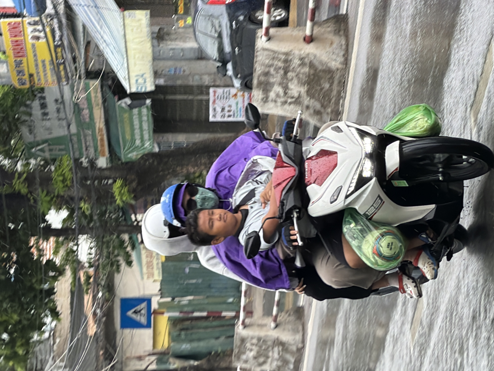 Three individuals are riding on a white motorcycle, moving from left to right on a wet street. The person in the front is a young boy wearing a gray shirt, white wristbands, and red and white Crocs. He is looking forward. Behind him, an adult wearing a purple jacket, blue helmet, and patterned face mask is riding as the second passenger, also looking forward. The third passenger, in the back, is wearing a white hoodie with the hood up and a white helmet, and is looking down. Two bright green plastic bags are attached to the sides of the motorcycle, one containing what appears to be a soccer ball. The environment appears to be a city street in Nha Trang, Vietnam, with buildings, signage, trees, and a black car visible in the background. The street is wet, suggesting recent rain or ongoing precipitation.