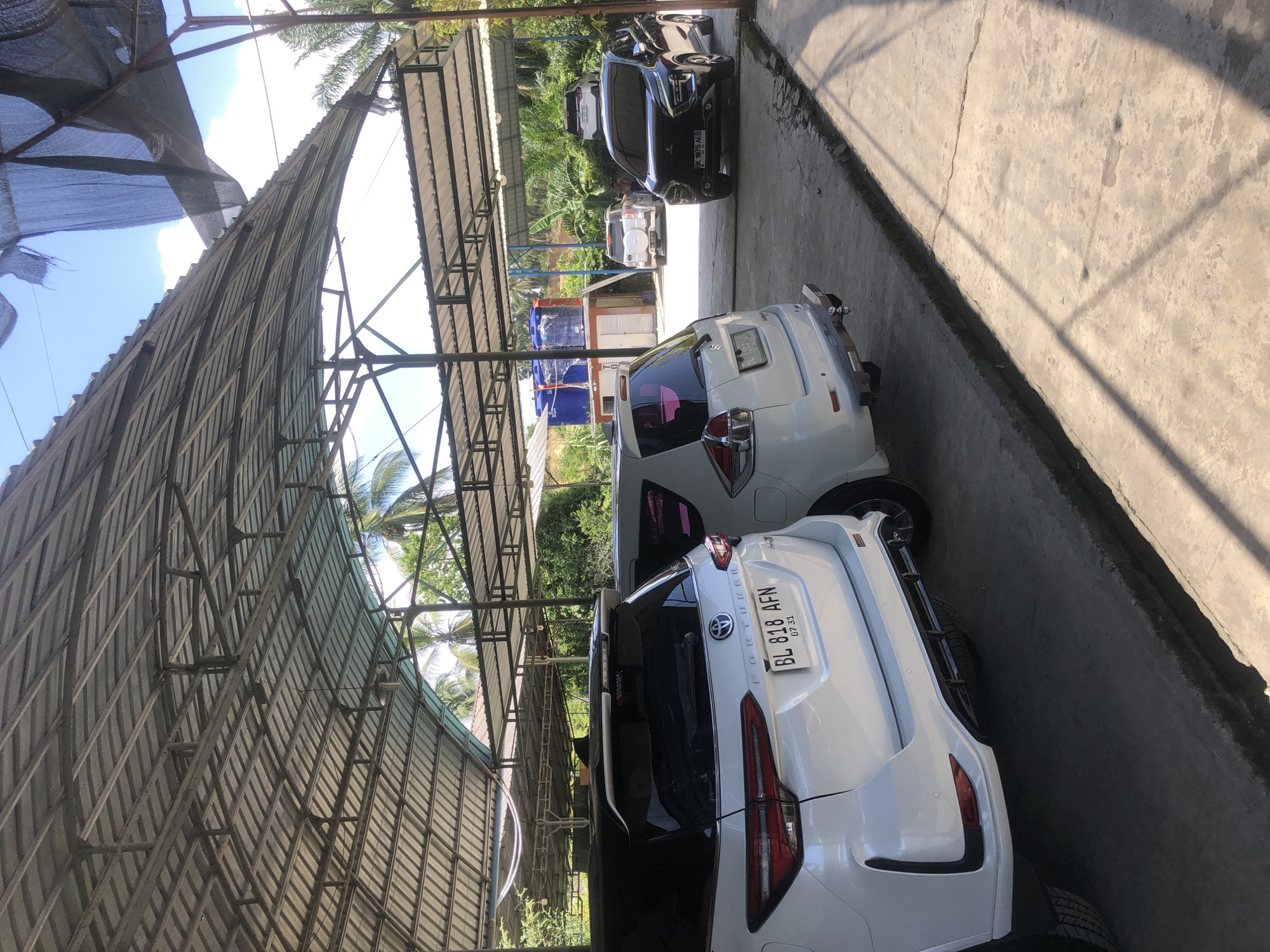 A covered outdoor parking area is shown with several vehicles parked under a corrugated metal roof supported by metal beams. In the foreground, a white Toyota Fortuner is parked on the left, and a white Suzuki Ertiga is parked to its right, partially obscured. Further back, a black Mitsubishi Xpander is parked on the right, facing left. Behind the Xpander, a white SUV is parked facing the camera. In the middle ground, a small building with blue water tanks on its roof is visible. The scene appears to be in a semi-rural or industrial area, with lush green vegetation and palm trees visible in the background. The ground is paved with concrete, and shadows from the roof and trees are cast across the surface. The lighting suggests it is daytime, with a bright sky visible through the gaps in the roof. The location is identified as Teupinbatee, Indonesia.
