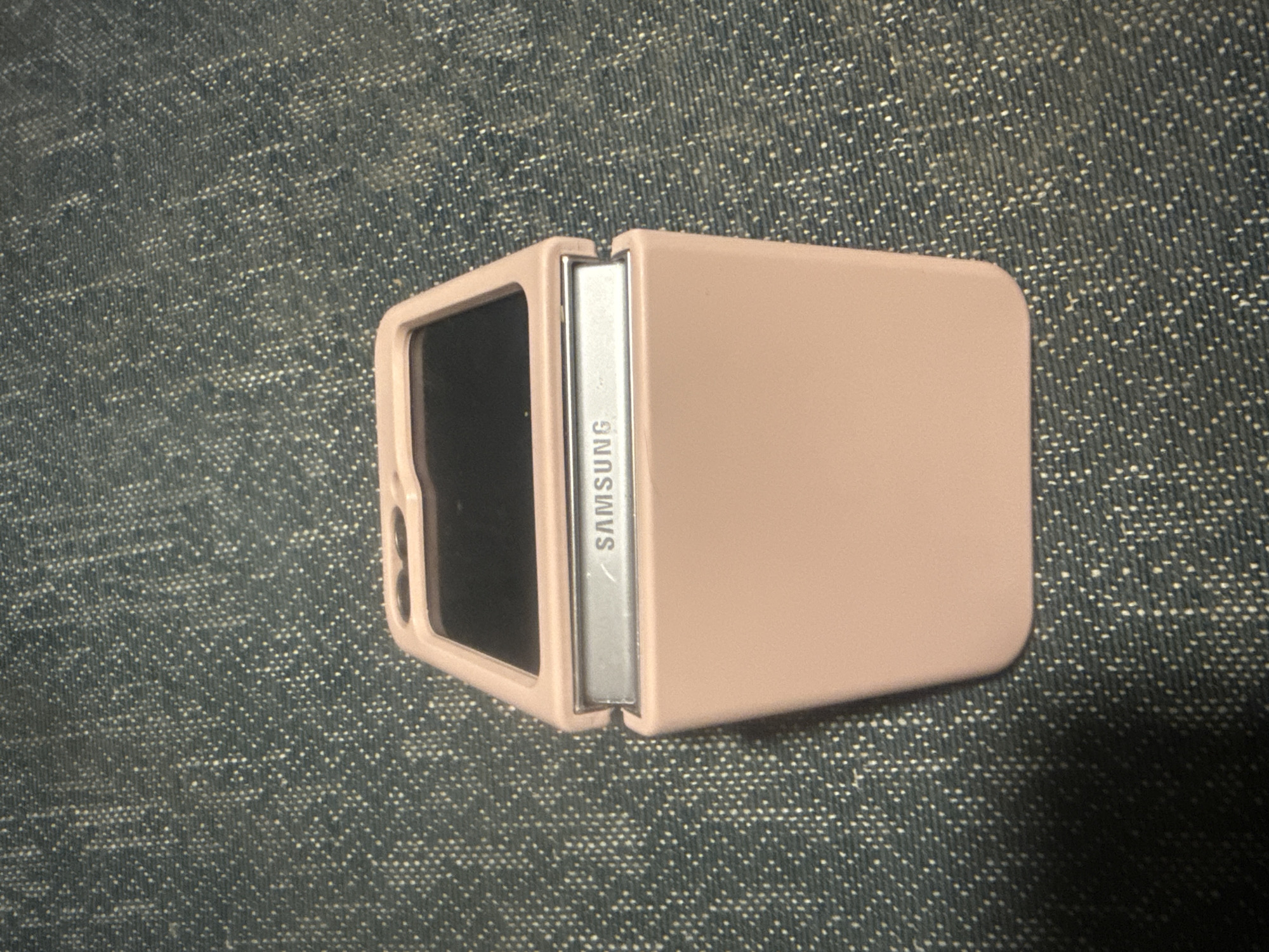 A pink Samsung flip phone case is open and lying flat on a dark blue denim-like surface. The phone's hinge is visible and has "SAMSUNG" embossed on a silver metallic strip. The top section of the case is angled upwards, revealing a dark screen. The background is textured with a pattern of small white speckles. The scene is lit from above, casting a subtle shadow to the left of the phone case. There are no people or other objects present. The image was taken indoors, with the background suggesting a domestic or casual setting. It is not possible to determine the exact location within Herzliya, Israel from the visual information provided.