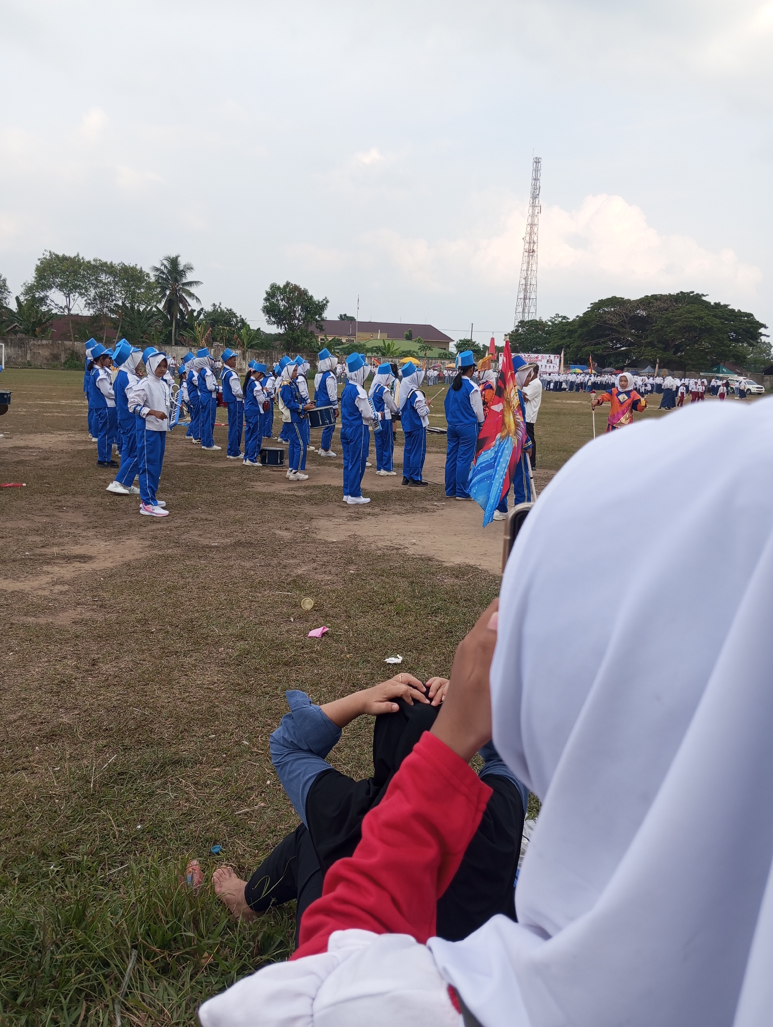 The image captures an outdoor event, likely a parade or performance, taking place in Candirejo, Indonesia. In the background, a marching band is lined up on a dirt field, with members wearing blue and white uniforms and blue hats. Some individuals are holding musical instruments like drums. Several flags are visible, including a large colorful one. Beyond the field, there are buildings, trees, and a tall telecommunications tower. In the foreground, two people are seated on the grass, facing away from the camera, with one person wearing a white headscarf and a red shirt. The overall scene suggests a community gathering with a focus on the marching band's performance.