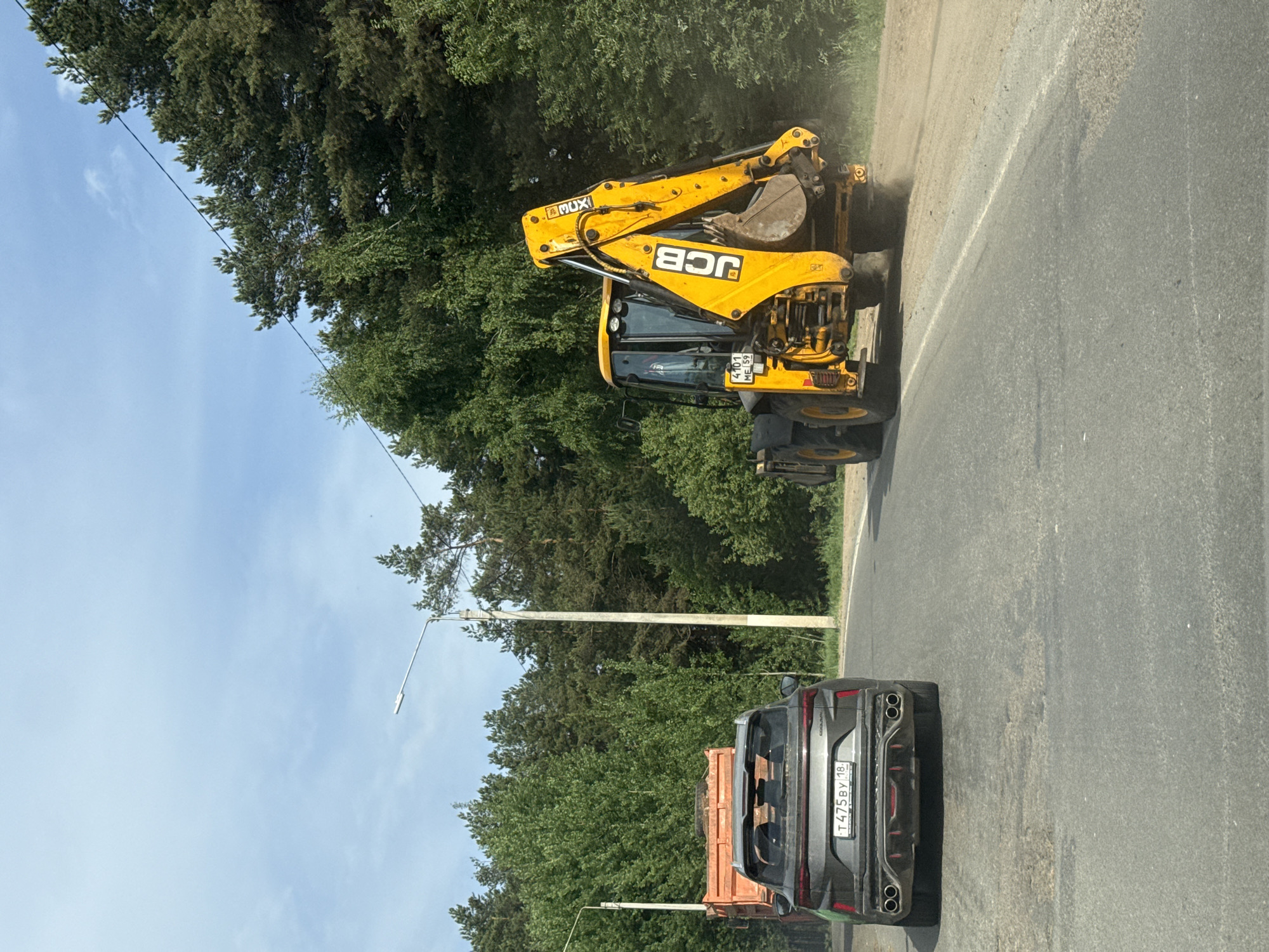 The image shows a JCB backhoe loader parked on the side of a road in Chaikovsky, Russia. Behind the loader is a gray car with a license plate that reads "T475BY 18". The surrounding environment includes trees, streetlights, and an overcast sky.