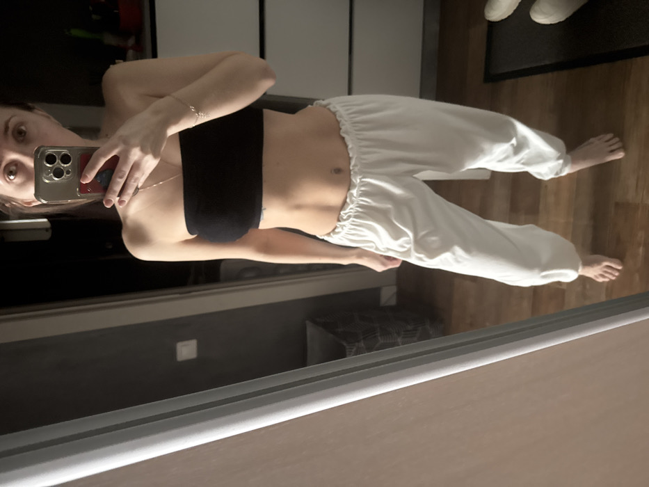 A person stands facing a mirror, holding a smartphone to take a selfie. The individual is wearing a black crop top and loose-fitting white pants with elastic at the waist and ankles. Bare feet are visible on a hardwood floor. Behind the person, there is a wardrobe or cabinet, and a dark, possibly carpeted area is visible in the reflection of the mirror. A light switch can be seen on the wall.