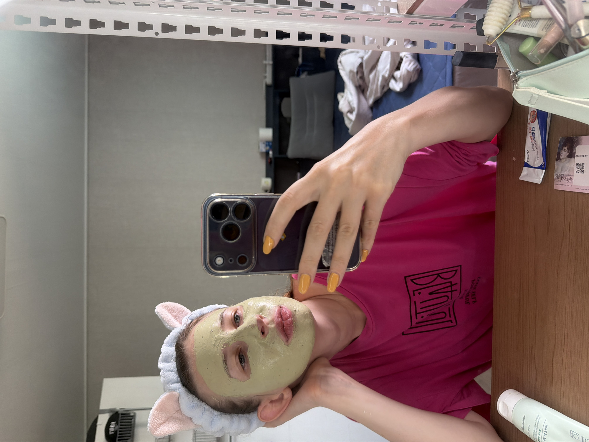 A young Caucasian woman with a green face mask and a pink cat-ear headband is taking a selfie in a mirror. She is wearing a bright pink t-shirt. Her left hand is resting on her cheek, and she is holding a smartphone in her right hand. The background shows a cluttered room with shelves, a bed, and some personal items. The scene appears to be in a residential setting, possibly an apartment, in Dongducheon-si, South Korea.