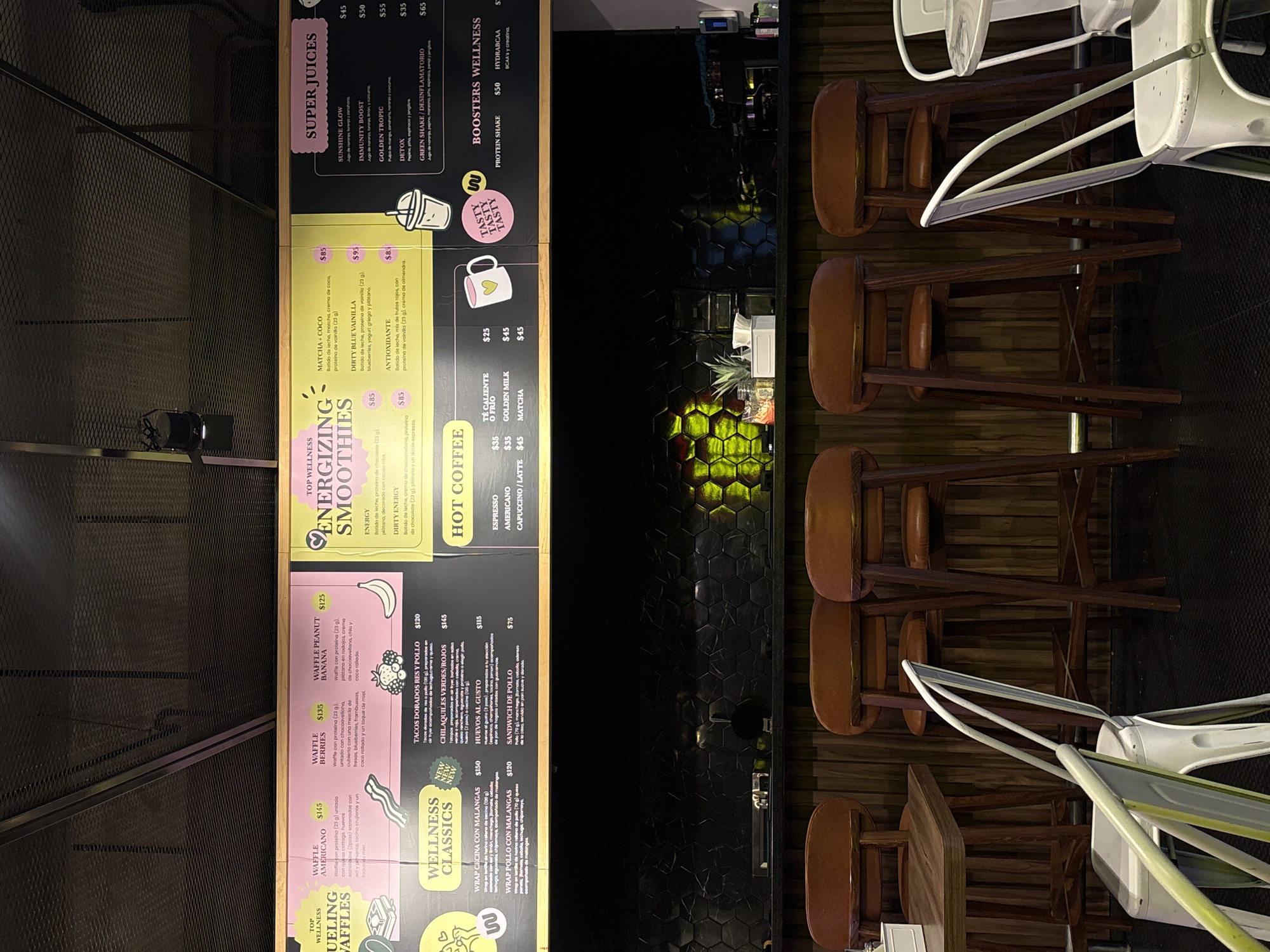 The image shows the menu of a food establishment. The menu lists several breakfast, smoothie, juice, and coffee options. The food establishment seems to be located in San Bernardino Tlaxcalancingo, Mexico. Additionally, the image shows a counter area with several high-top chairs, potentially indicating a dining space. The menu board itself is split into sections, each highlighting different categories of food and drinks offered by the establishment.