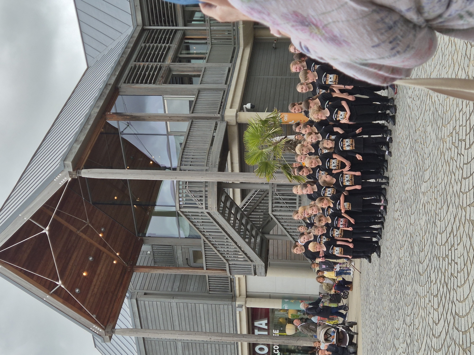 A group of approximately 25 children and teenagers, mostly girls, are arranged in several rows, standing formally on a patterned stone plaza. They are uniformly dressed in black t-shirts with white lettering that reads "ROCK YOU" and "young@sport," facing towards the right side of the frame. In the extreme right foreground, the out-of-focus shoulder and arm of an individual holding a phone or camera are visible, indicating they are photographing the group.
The backdrop is a modern, multi-story building identified as the Arc in Bury St Edmunds, West Suffolk, United Kingdom. The building features an exterior clad with light grey wooden slats, large glass windows, and a prominent, expansive wooden canopy over an entrance, supported by a triangular steel framework and featuring recessed lighting. A substantial external wooden staircase with railings ascends to an upper level on the left side of the building. To the far left, a "Costa" coffee shop occupies the ground floor, with pedestrians visible nearby. The plaza surface is paved with light grey, irregularly shaped stones laid in radiating and concentric patterns. A small palm tree is situated to the left of the children. The sky is overcast.