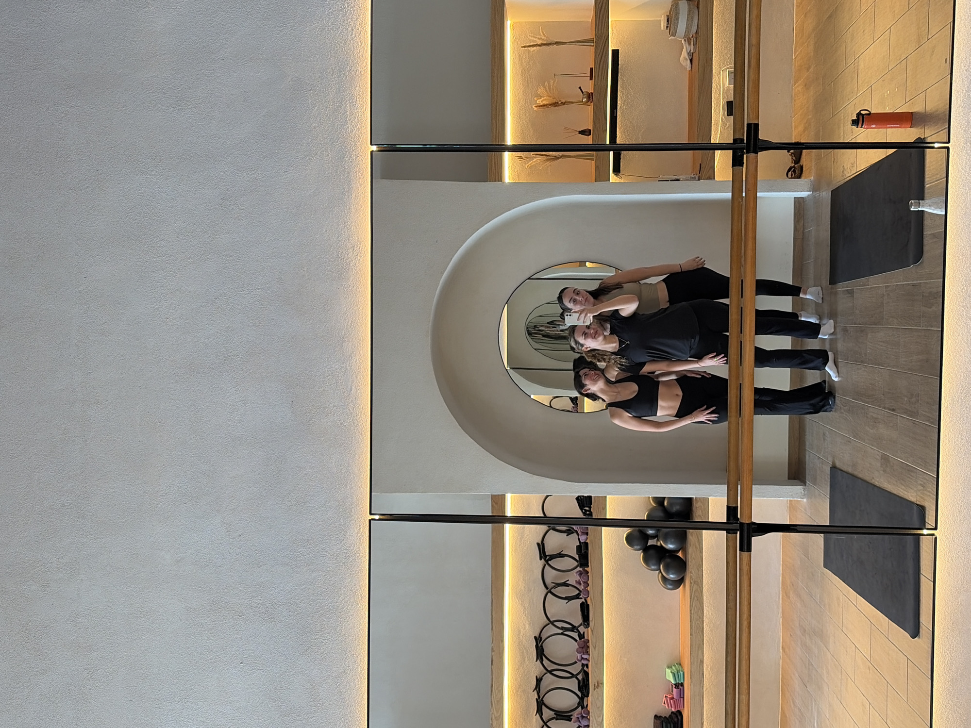 The image captures an indoor scene within a modern fitness or exercise studio, specifically reflecting three women through a series of large, wall-mounted mirrors. The setting appears to be a Pilates or barre studio, given the visible exercise barre and athletic wear. The studio is located in San Bernardino Tlaxcalancingo, Mexico.
In the central reflection, two women are the primary subjects. One woman, with light brown hair and wearing a black sports bra and pants, is holding a smartphone and appears to be taking a selfie. Behind her, another woman with dark hair, dressed in a beige sports top and black leggings, smiles towards the camera. A third woman, also in black athletic attire, is partially visible to the left in the reflection.
The studio's interior features light-colored, textured walls with subtle, warm recessed strip lighting running along vertical edges and illuminating shelves. The floor is covered with light brown, rectangular tiles. A wooden exercise barre is prominently positioned across the mid-ground of the reflected image. Exercise equipment such as black stability balls and resistance bands are visible on racks and shelves. Decorative elements like dried pampas grass are placed on wooden shelves, also highlighted by warm lighting. A distinctive feature is an arched alcove containing a circular mirror. On the tiled floor, a red water bottle with the brand "Pioneer" visible on it, rests near a black exercise mat. The overall atmosphere is bright, clean, and modern.
