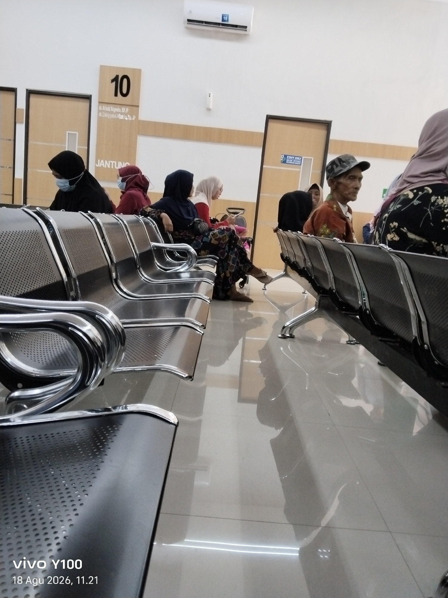 The image depicts a waiting area, likely in a medical facility, with several individuals seated on metallic benches. The environment appears to be indoors, with tiled flooring that reflects the overhead lighting. To the left of the frame, a sign indicates "JANTUNG" and the number "10", suggesting a cardiology department or a specific examination room. Several people are wearing headscarves and face masks, common in public spaces in Indonesia. One man, in the center, is wearing a patterned shirt and a cap. The overall impression is one of a calm, albeit somewhat sterile, waiting space.