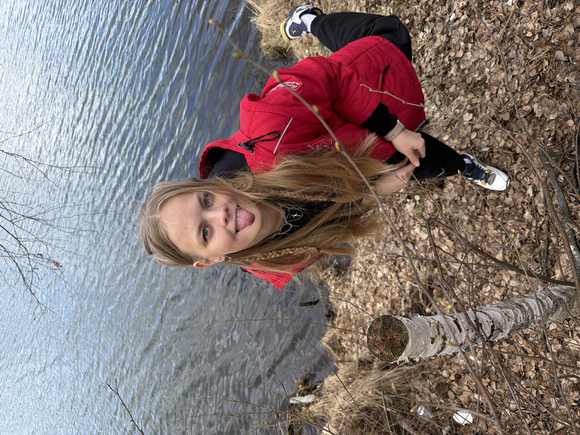 An outdoor portrait shot in Novyi, Russia, features a young person posing playfully by a body of water. The subject has long, light brown hair and is looking directly at the camera with their tongue slightly extended, conveying a lighthearted and casual mood. They are dressed in a bright red hooded jacket, a black long-sleeved shirt, and black pants. A watch is visible on their left wrist, and a necklace with a cross pendant is worn around their neck. On the jacket's right chest area, a white patch with partially obscured black text is visible, possibly including words like "NEW YORK" or "PLAYER." Their footwear consists of white sneakers with yellow and black accents, featuring a recognizable athletic shoe brand logo.
The person is positioned near the edge of what appears to be a lake or river, with their right leg bent and resting on the ground. The immediate foreground and midground on the right are covered with dry, fallen brown leaves and bare, thin tree branches with visible small buds, suggesting a late winter or early spring season. To the right, a segment of a white-barked log, possibly birch, lies horizontally on the leaf-covered ground. The body of water, which dominates the left side of the frame, exhibits ripples across its surface, reflecting the bright sky, indicating daytime. The natural setting, with water, trees, and fallen leaves, creates a rustic and serene backdrop for the playful subject.