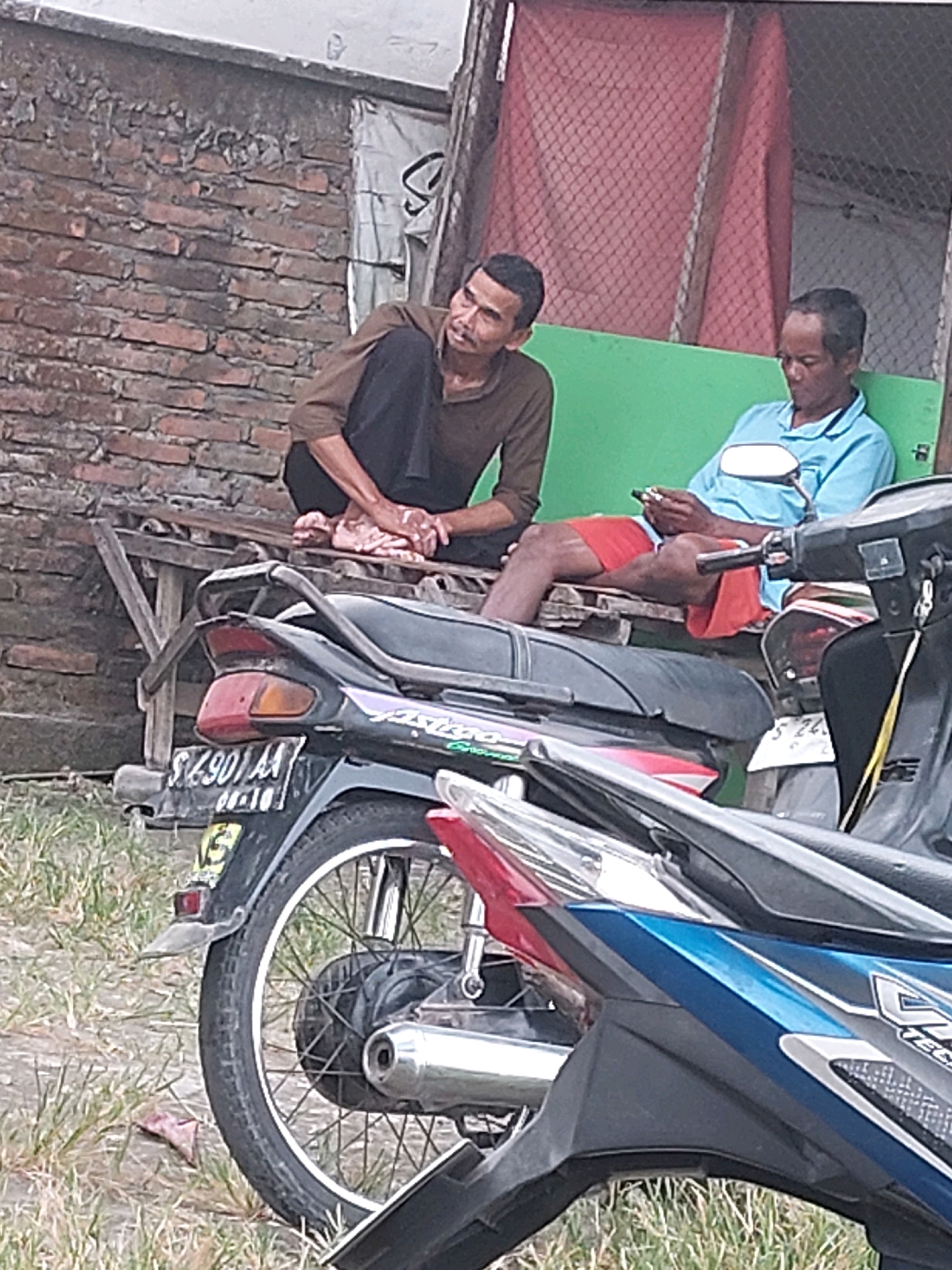 Two men are seated on a wooden bench outdoors. The man on the left wears a brown long-sleeved shirt and dark pants, and is leaning back with his legs crossed and feet resting on the bench. He appears to be smoking. The man on the right wears a light blue collared shirt and red shorts, and is looking down at his mobile phone. They are seated in front of a brick wall and a green structure, with a motorcycle parked in front of them. The ground is covered in dry grass and dirt. The scene is likely located in SARANGAN, Indonesia.