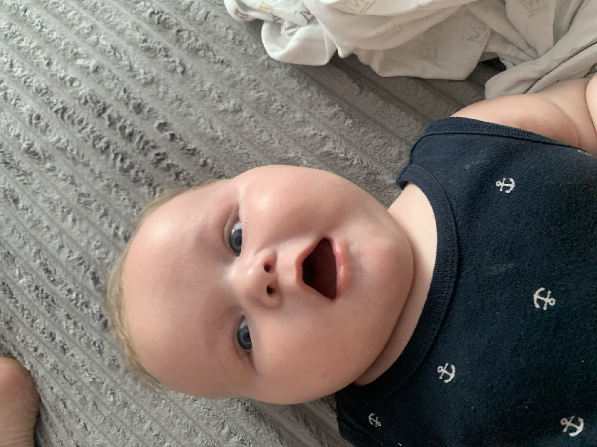 This media file features a close-up, overhead shot of a baby lying on a grey, textured surface. The baby is wearing a dark blue onesie with a pattern of white anchors. The baby's face is the central focus, with wide, curious blue eyes and an open mouth, suggesting surprise or excitement. Their cheeks are full and rosy, and a few wisps of fair hair are visible. To the upper right of the image, a crumpled white blanket with a subtle pattern is visible, possibly indicating a home setting. The lighting appears to be natural, suggesting it might be daytime. The overall mood is one of innocence and wonder. No text is visible in the image.