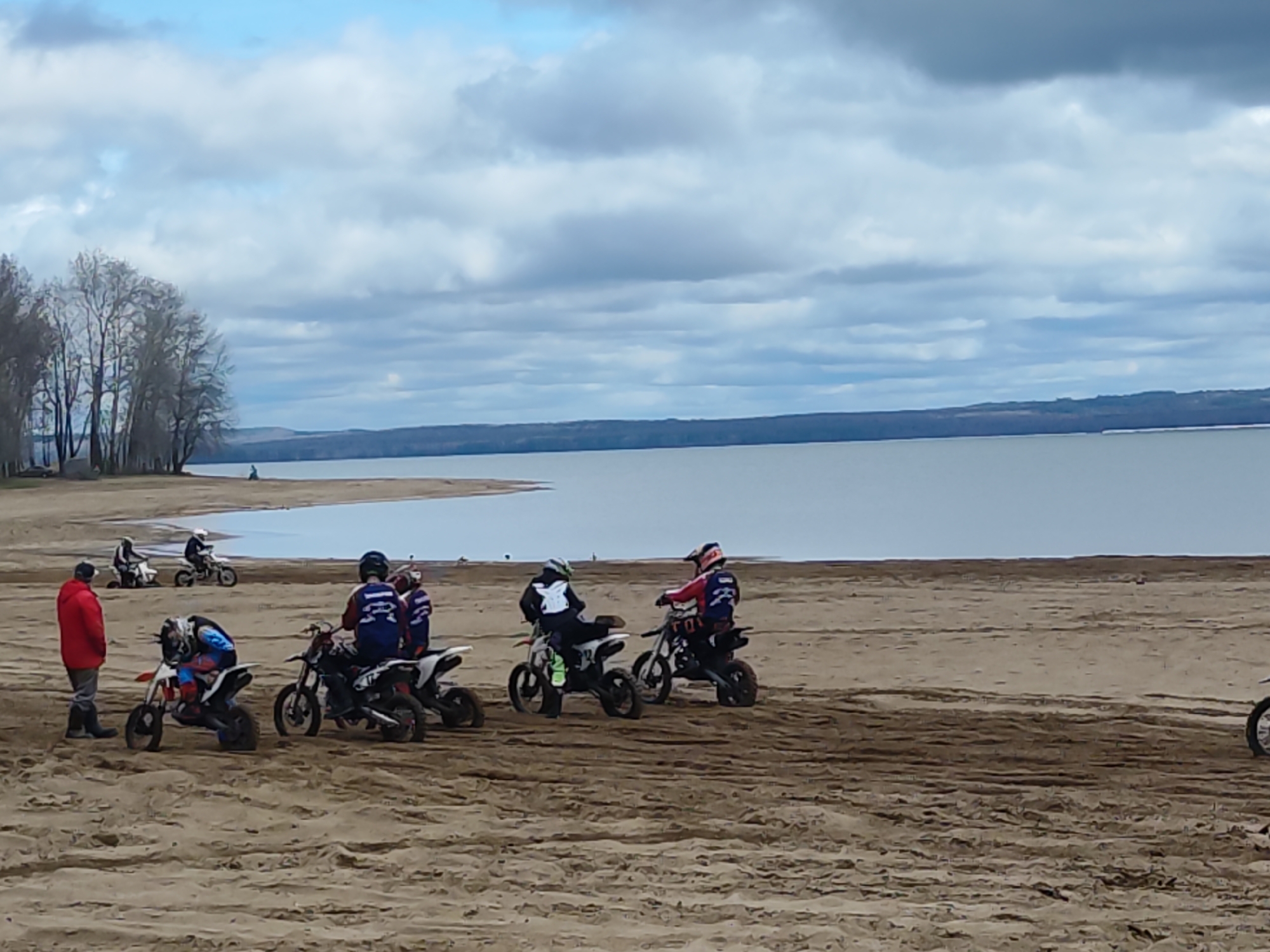 The image depicts a group of people on motorcycles at a sandy beach near a body of water. There are at least six motorcycles visible, with some riders seated on them. A man in a red jacket stands near the motorcycles. In the background, there is a lake or river, a strip of land, and a cloudy sky. The location is Chaikovsky, Russia.