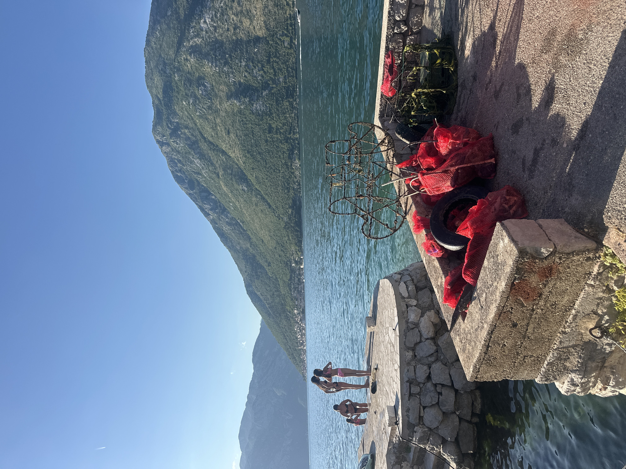 The image captures a sunny outdoor scene in Prčanj, Montenegro, featuring a body of water, mountains, and a dock with people and fishing equipment.
In the foreground, a concrete and stone dock extends into the water. On the main concrete section of the dock, various items are stored: multiple rusted metal structures resembling old fishing traps or pots, several red mesh bags (some appearing full), a few black rubber tires, and a green net or basket. Long, distinct shadows are cast across the dock surface, indicating bright sunlight from the left.
To the left of the concrete dock, a circular stone platform serves as a swimming area. Four individuals, appearing to be children or young adults, are present on or near this platform. One child in dark swimwear is partially submerged in the water by the platform's edge. Another individual in dark swimwear is emerging from the water onto the platform. Two other individuals, one in dark swimwear and one in a pink bikini, stand on the platform, facing the water.
The midground features a calm, blue-green body of water (the Bay of Kotor). A white speedboat is visible in the middle distance, moving rightward and leaving a wake.
The background is dominated by large, steep, green mountains, characteristic of the Bay of Kotor, rising directly from the water's edge. On the opposite shore, nestled at the base of these mountains, a cluster of buildings forms a village, with some structures appearing as traditional stone houses and potentially a church steeple. More mountains recede into the distance on both sides of the bay.
The sky is clear and bright blue, occupying the upper portion of the frame. A faint white contrail from an aircraft is visible in the upper left quadrant of the sky.