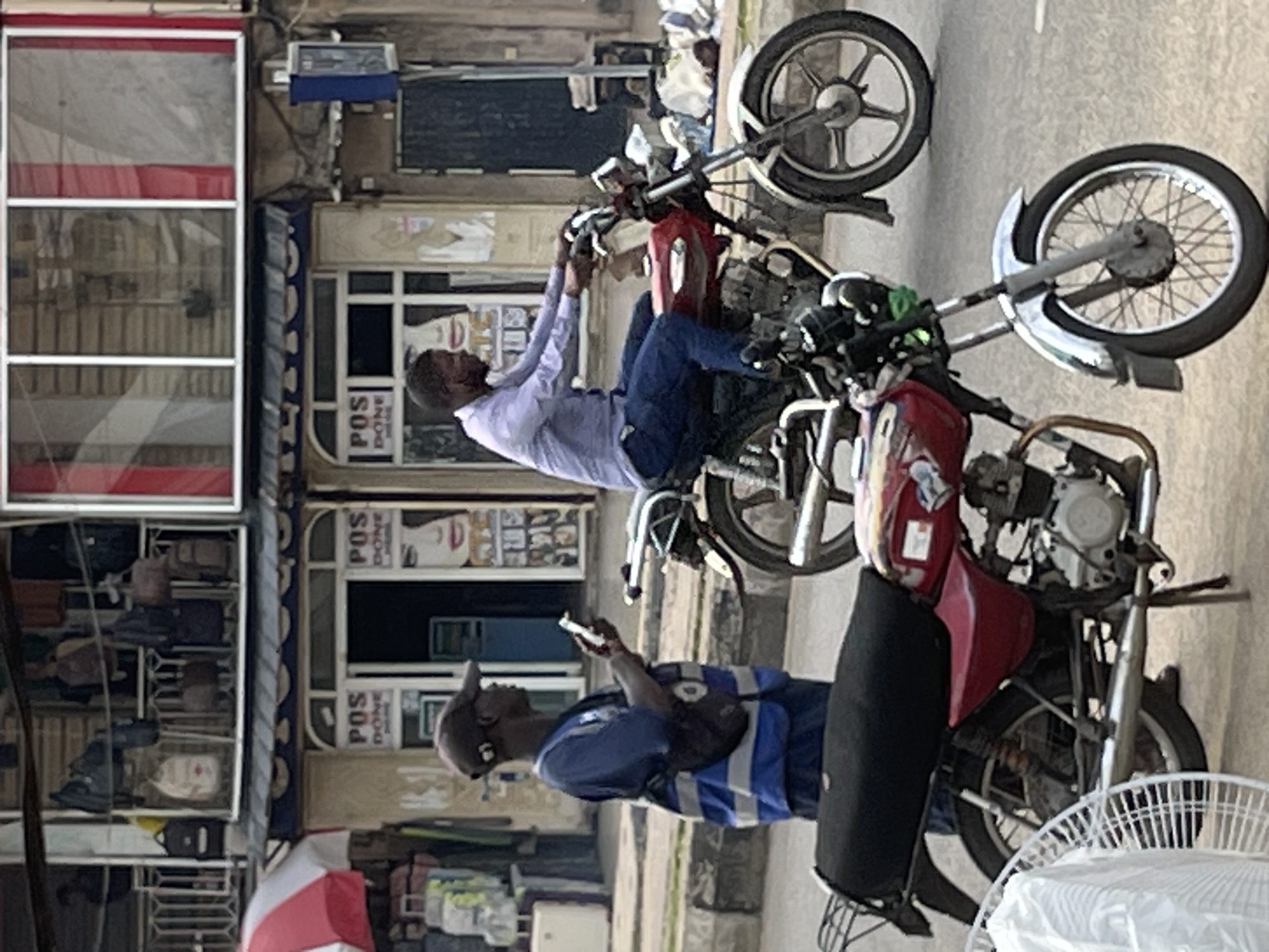 The image shows two men and two motorcycles on a street. The man in the foreground is wearing a blue vest over a blue shirt and a dark baseball cap. He is looking at his phone. The man behind him is wearing a light purple shirt and blue pants. He is sitting on a red motorcycle and appears to be holding the handlebars. The second motorcycle is partially visible behind the man in the red motorcycle. Both motorcycles appear to be in working order.
In the background, there are buildings with windows and signs. One sign appears to say "POS DONE." There are also some items displayed in a window that look like bags. A red and white umbrella is visible in the lower left portion of the image.
The scene appears to be taking place during the daytime. The lighting suggests it could be a bright day. The environment seems to be an urban or semi-urban area. The overall impression is of a typical street scene with people and vehicles. The people's expressions are neutral, and the activity is mundane.