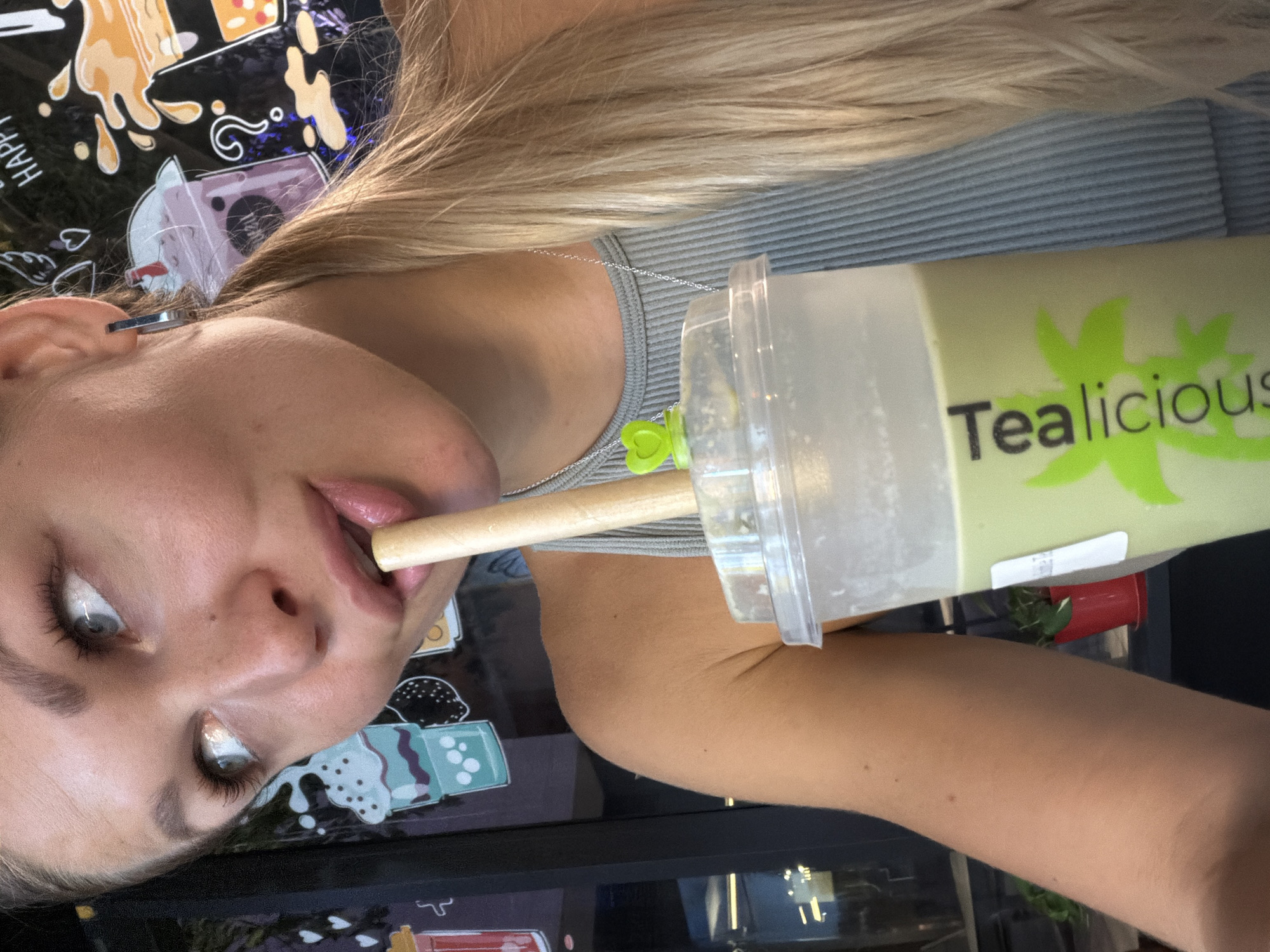 This close-up, angled photograph captures a person in the act of drinking a beverage from a large cup. The subject, a person with long blonde hair, is partially visible, showing her left ear adorned with a silver hoop earring, her eye looking slightly upwards, and her lips around a wooden straw. She is wearing a grey ribbed top and a silver chain necklace with a small green heart-shaped charm rests near her collarbone.
The prominent object is a clear plastic cup filled with a light green beverage, likely a bubble tea. The cup features a bright green logo with the word "Tealicious" in a distinctive font, accompanied by a leaf-like symbol above the 'i'. A thick wooden straw extends from the cup to the person's mouth.
The background is a dark, reflective surface, possibly a table or counter, decorated with colorful illustrations of various bubble tea cups and splashes. Visible partial text includes "HAPPY" and a question mark. Further in the background, a red cup and some greenery are subtly discernible. The lighting suggests it is daytime. The setting appears to be a casual cafe or bubble tea shop, consistent with a "Tealicious" establishment in Dubai, United Arab Emirates. The image conveys a casual moment of refreshment.