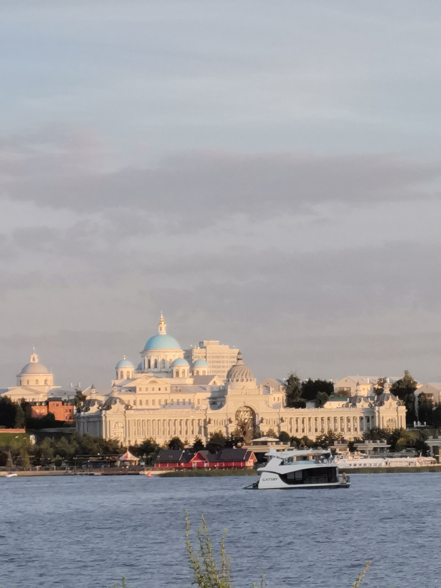 The image depicts a wide, calm body of water in the foreground, likely a river or lake. A modern white yacht with the name "GATSBY" visible on its side is cruising on the water. In the middle ground, a complex of white buildings with blue domes and classical architectural elements lines the shore, with trees interspersed. The sky above is a soft gradient of light blue, with subtle cloud formations. The overall scene suggests a peaceful waterfront view in the city of Kazan, Russia, likely the Kazan Kremlin or a nearby area. The lighting indicates it may be late afternoon or early evening.