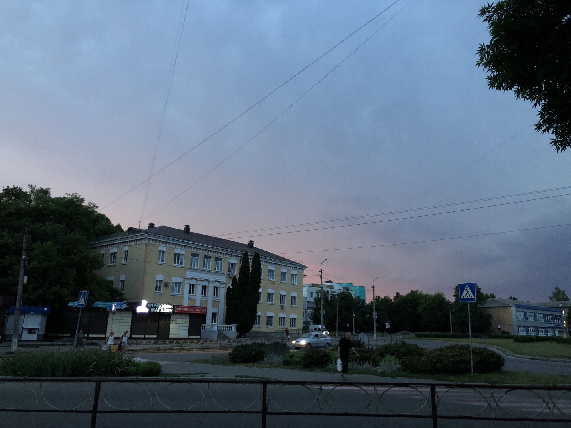 The image depicts a street scene at dusk in an urban environment. A multi-story yellow building with white trim and numerous windows is a prominent feature on the left side of the frame. Several smaller structures and a larger blue building are visible further down the street. A pedestrian crossing sign is displayed prominently on a pole. The sky is a soft gradient of purple and blue, indicative of twilight, with power lines stretching across it. Trees and greenery line the street, and a few individuals are walking. A car is driving on the road. The scene is framed by a metal railing in the foreground.