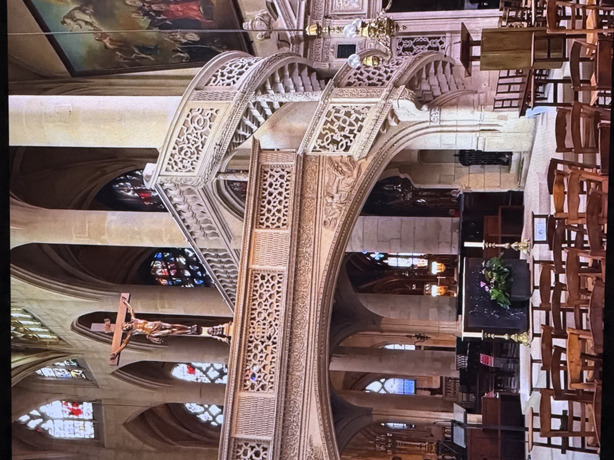 This media file shows the interior of a church. The scene is a grand, ornate interior with high ceilings, arched windows, and intricate architectural details. Dominating the foreground is a large, carved wooden crucifix depicting Jesus Christ. Behind the crucifix, stained glass windows are visible, casting colorful light. The architecture suggests a European style, possibly Gothic or Romanesque, with decorative stonework and soaring columns. In the background, rows of wooden chairs are arranged, indicating this is a place of worship.
The lighting suggests it is daytime, with light filtering through the stained glass windows. There are no people clearly visible in the frame, but the presence of the chairs and the crucifix implies a religious setting. The overall atmosphere is one of solemnity and reverence. There is no visible text in the image. The location context provided (Dubai, United Arab Emirates) appears to be incongruent with the visual cues of the image, which strongly suggests a historic European church.