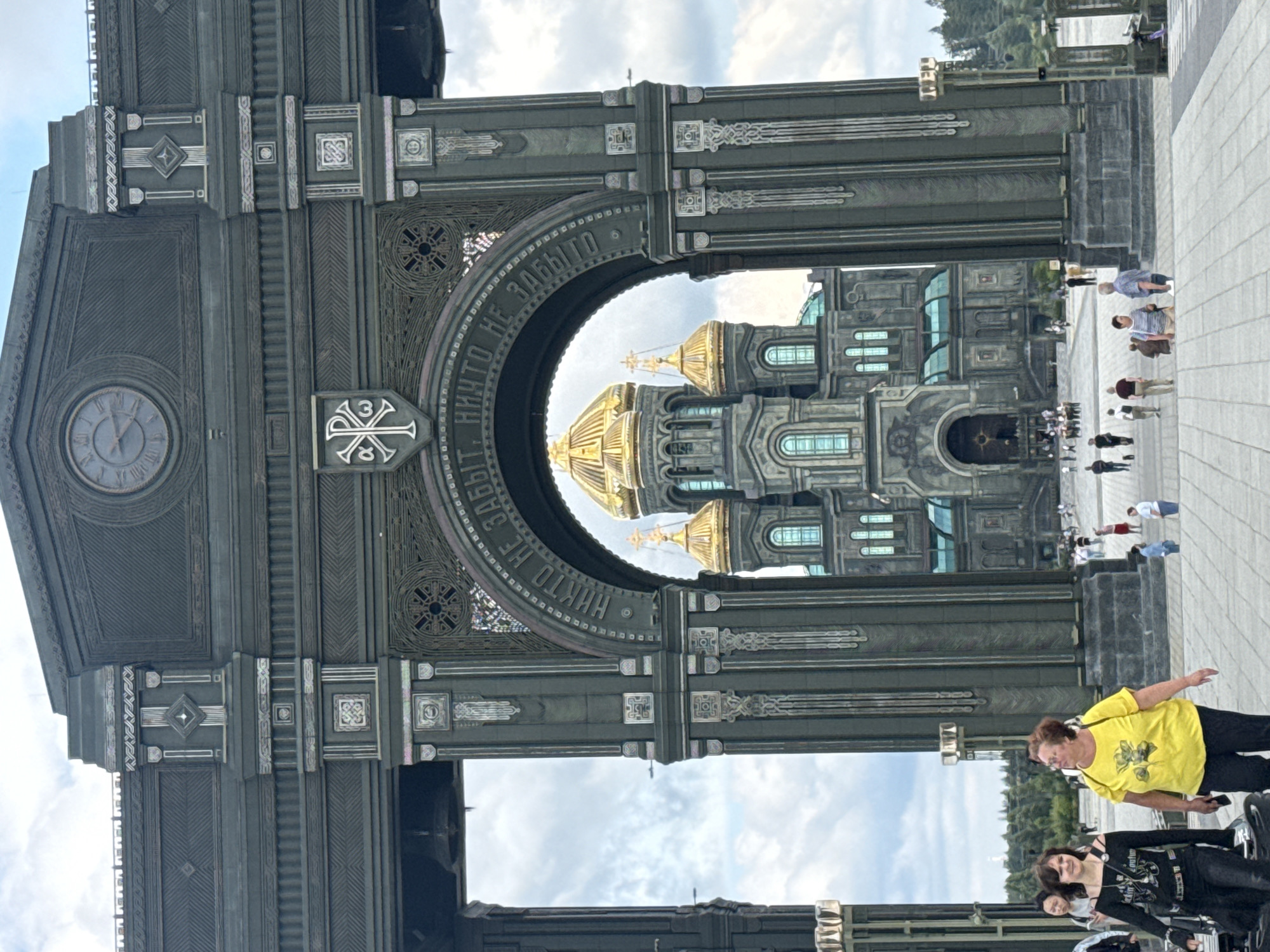 This image shows the Patriarchal Cathedral of the Russian Armed Forces in Kubinka, Moscow Oblast, Russia, viewed through a large, ornate archway. The cathedral, with its golden domes and grey facade, is the central subject. The archway is made of dark metal and features intricate carvings and inscriptions. Several people are visible in the foreground and on the plaza in front of the cathedral, appearing to be tourists or visitors. Some are walking, while others are standing and observing the architecture. The sky is partly cloudy. The scene appears to be a sunny day with moderate foot traffic.
