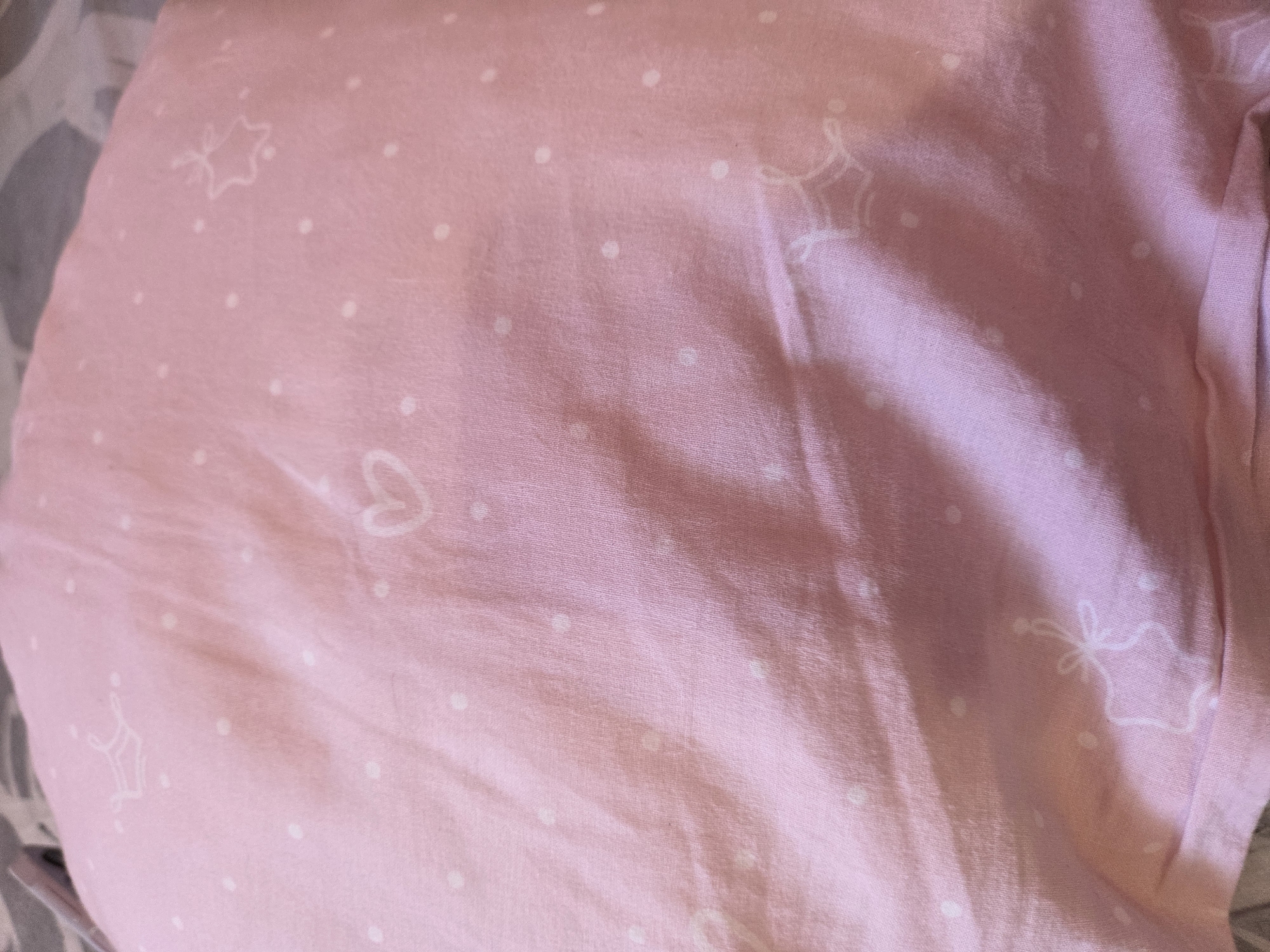 The image presents a close-up view of a pink fabric, likely a pillowcase or bedding, in an indoor setting within Krastevich, Bulgaria. No individuals or specific activities are depicted.
The fabric is a soft, light pink hue with a visible weave and gentle creases and folds, suggesting a soft material such as cotton. It features a decorative pattern consisting of scattered small white polka dots. Interspersed among the dots are delicate white outline drawings. These include five-pointed stars, some of which are embellished with a small bow-like detail extending from one of their points. A single outline of a heart shape is also discernible towards the top-center of the frame.
The lighting is soft and diffused, casting subtle shadows within the fabric's folds and highlighting its contours. This suggests natural light, possibly from a window, rather than harsh artificial illumination. The overall aesthetic is one of softness, comfort, and a gentle or youthful theme, evoked by the pastel color and playful patterns. There is no visible text present in the image.