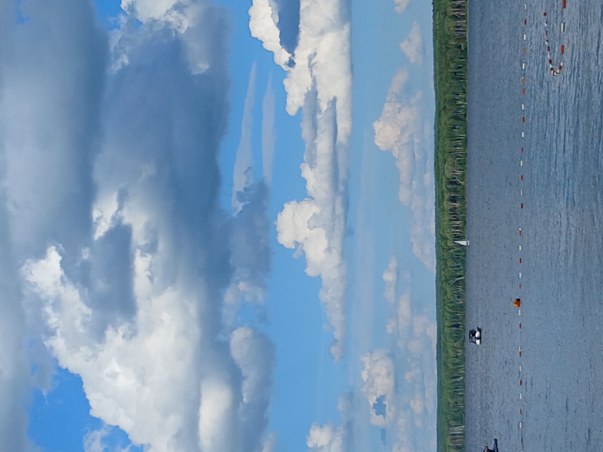 This is a vertical, outdoor, eye-level shot of a body of water. The water is a dull blue and appears to be a lake or calm sea, with ripples visible. A thin line of trees, likely a forest, runs along the far edge of the water, creating a dark green border against the sky. The sky is a vibrant blue, filled with a mix of large, fluffy white clouds and darker gray storm clouds. The clouds are dramatic and dynamic, suggesting a partly cloudy or changing weather condition.
In the water, several small buoys are visible, marking a boundary or a course. One orange buoy is particularly noticeable. A small white sailboat can be seen in the distance, sailing on the water. Closer to shore, a small black boat with two people in it is also present. The overall scene is peaceful and natural, with a sense of open space. The time of day appears to be daytime, given the bright sky and evident sunlight on the clouds and water. The weather is variable, with both sun and cloud cover. The location cues, such as the forest, the type of water, and the buoys, are consistent with a recreational body of water. No text is visible in the image.