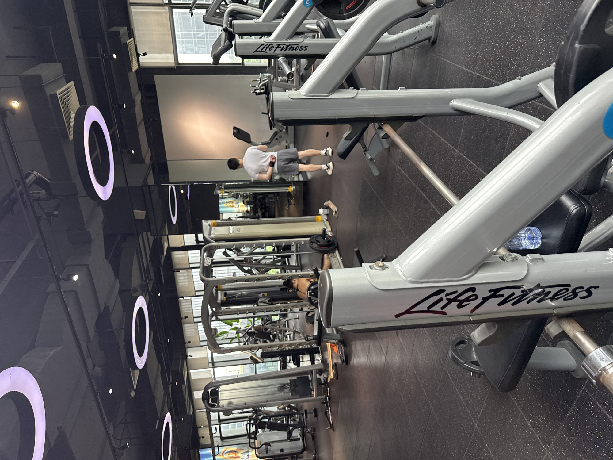 An interior shot captures a gym environment in Haizhu District, China. The scene is dominated by various exercise machines, primarily from the brand Life Fitness, with their branding visible on the equipment. In the foreground, a close-up view of a weight bench and its supporting structure is prominent. The flooring is a dark, speckled rubber matting.
In the mid-ground, a man wearing a white t-shirt and grey shorts stands facing away from the camera, appearing to be taking a break or waiting. Further in the background, another individual, a woman, is lying on a weight bench with weights positioned near her, suggesting she might be in the middle of a workout.
The gym ceiling features circular, illuminated light fixtures and exposed ductwork. Large windows line the back wall, allowing natural light to filter in and offering a glimpse of buildings and greenery outside, consistent with an urban setting. The overall impression is of a modern, well-equipped fitness facility.
