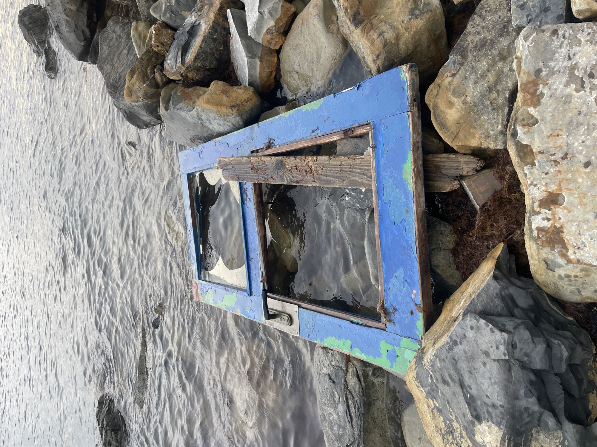 The image shows a discarded, weathered blue-painted window frame lying on the edge of water, possibly a shore. The frame is partially submerged in the water and surrounded by rocks and possibly algae. Some of the glass panes are broken, and a piece of wood is wedged within the frame. The water is rippled. The location is said to be Olginka, Russia. There are no people or actions visible in the image.