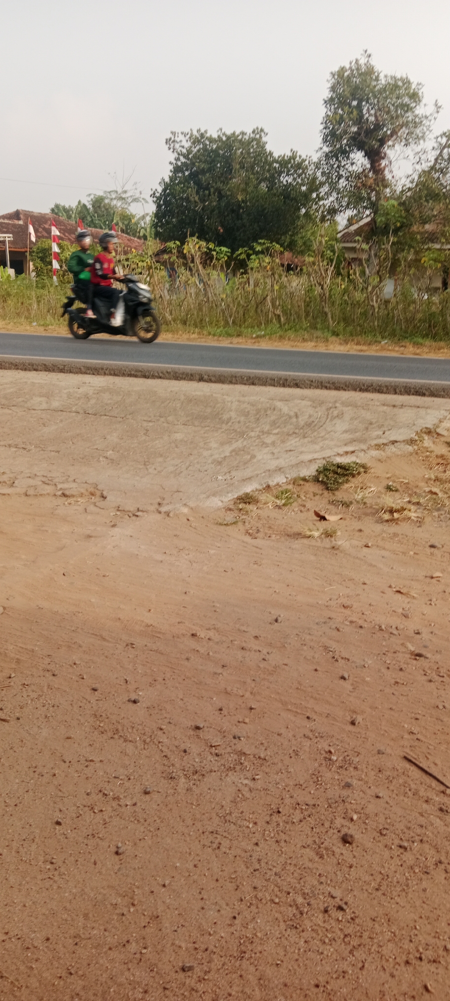 Two individuals, wearing helmets, are riding a black motorcycle on a paved road. The person in the front is wearing a red shirt and the person in the back is wearing a green jacket. They are moving from left to right across the image. The road is bordered by dry grass and trees on the right, and a small building with red and white flags on the left. The ground in the foreground is dry, sandy soil with some scattered pebbles. The sky is overcast. The scene appears to be in a rural or semi-rural area, likely in Gondang Rejo, Indonesia.