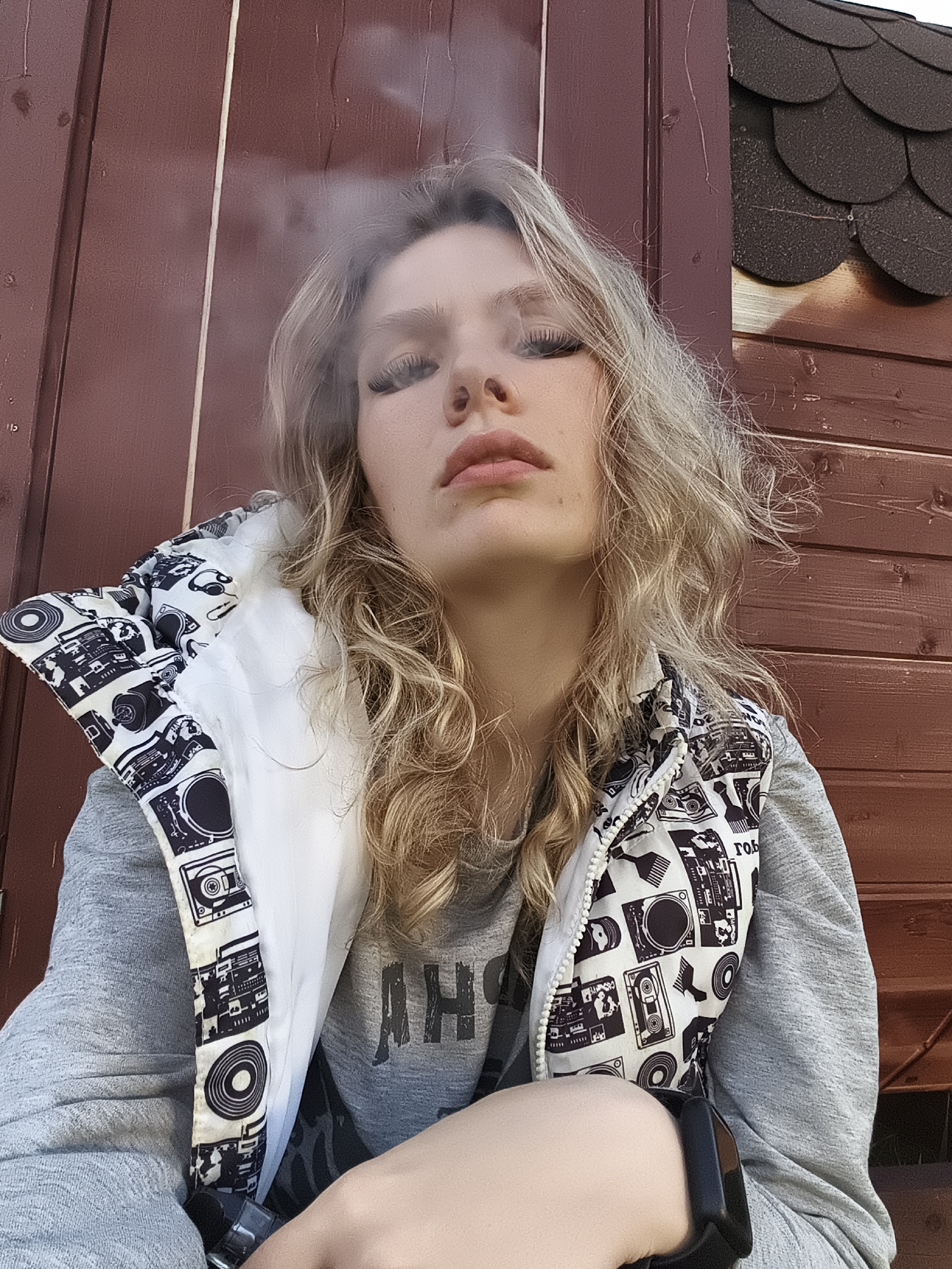 A young woman with long, wavy blonde hair is depicted in a close-up portrait, looking directly at the camera with a confident and direct gaze. She wears a grey long-sleeved shirt with partially visible text, possibly "АНЬ" in Cyrillic, underneath a distinctive white and black patterned sleeveless puffer vest. The vest's pattern features various vintage audio equipment, including boomboxes, cassette tapes, vinyl records, and headphones, with some small, illegible text like "ROB" visible. A black smartwatch or fitness tracker is worn on her right wrist.
Wisps of white smoke or vapor are visible above her mouth, suggesting she may be exhaling vapor from an e-cigarette or her breath in cool air. The background consists of a reddish-brown wooden wall made of horizontal planks, likely part of a building or shed. A dark, shingled roof is partially visible in the upper right corner. The scene is set outdoors in daytime, with lighting suggesting morning or afternoon. The clothing and visible vapor imply cooler weather conditions. This photograph was taken in Русский Вожой, Russia.