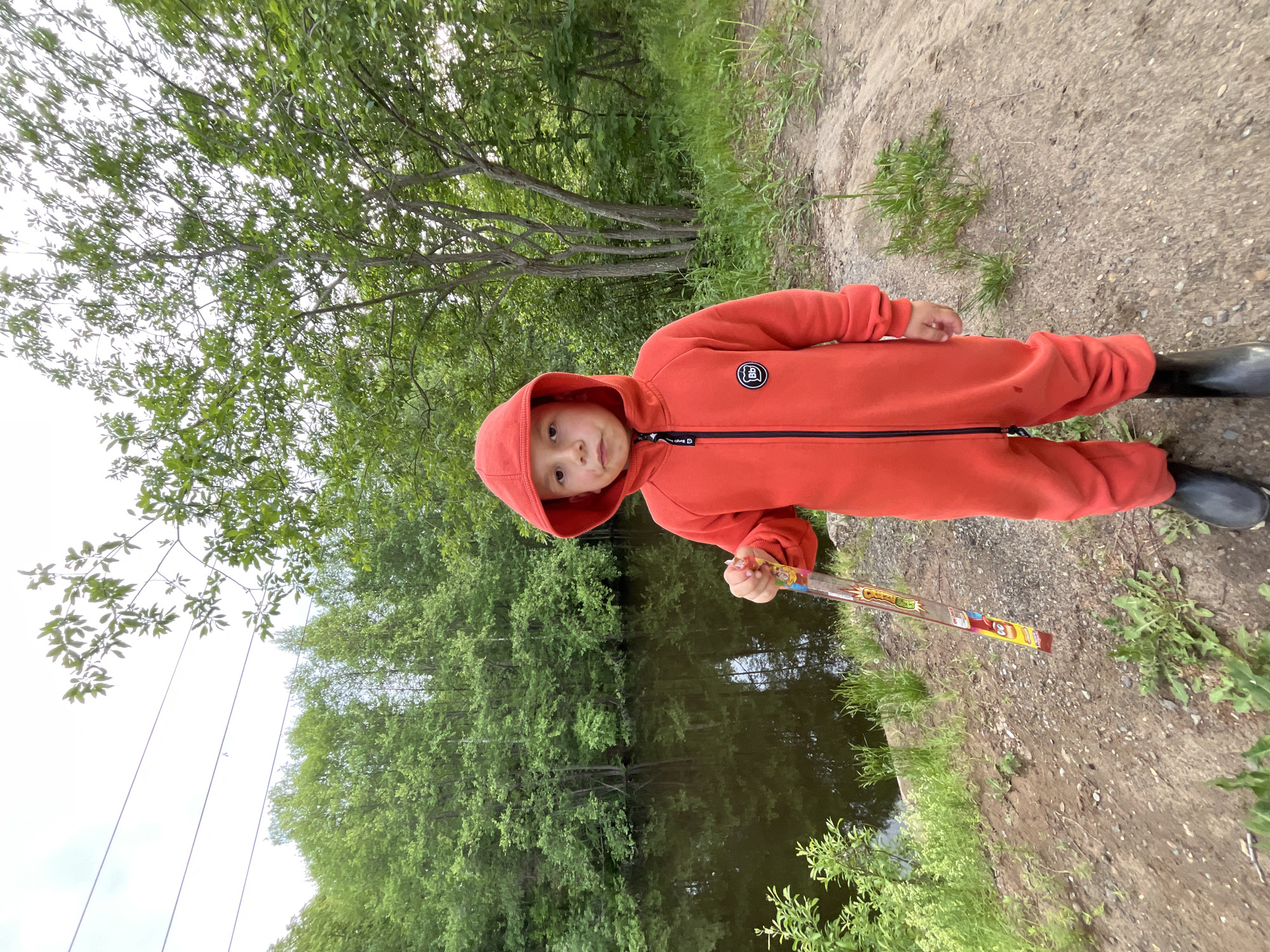 A young child in an orange hooded jumpsuit and black boots stands on a dirt path next to a body of water. The child is holding a long, colorful object resembling a candy stick or toy. Behind the child, trees line the water's edge, suggesting a natural or park setting. The location is near a pond, near the city of Chaikovsky, Russia.