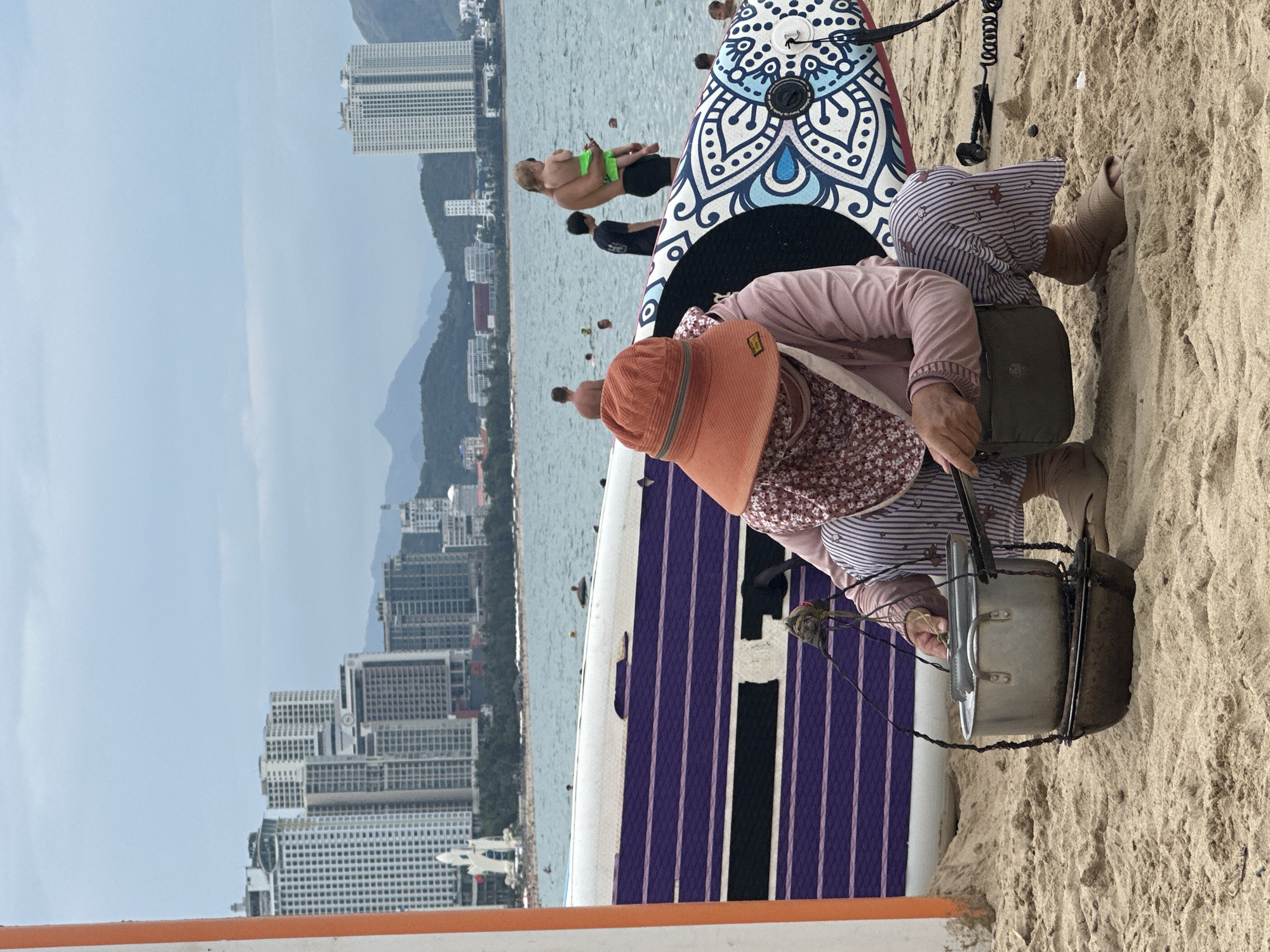 This image depicts a lively beach scene in Nha Trang, Vietnam, under a bright, clear sky.
In the foreground, an elderly person is seated on the sandy beach. They are wearing a wide-brimmed orange sun hat, a long-sleeved light pink top, and distinctive striped pants. They appear to be attending to a metal pot or cooking container, which is suspended over a small stand directly on the sand. A small, dark bag or purse is visible next to them. Two paddleboards are propped up on the sand nearby; one features an intricate blue, white, and black mandala-like pattern, while the other has purple and black stripes. A coiled leash is attached to one of the boards.
In the mid-ground, the tranquil blue ocean stretches out, where several people are engaged in activities. Some individuals are swimming or wading, and a person in green shorts is seen carrying or assisting another person in the water.
The background showcases a dense urban landscape, typical of a coastal city. Numerous tall, modern high-rise buildings line the shore, indicating a developed area. Beyond the cityscape, hazy mountains are visible in the distance, completing a panoramic view that combines natural beauty with urban development. The scene suggests a busy day at the beach, featuring both recreational activities and local commerce. No clearly legible text is visible in the image.