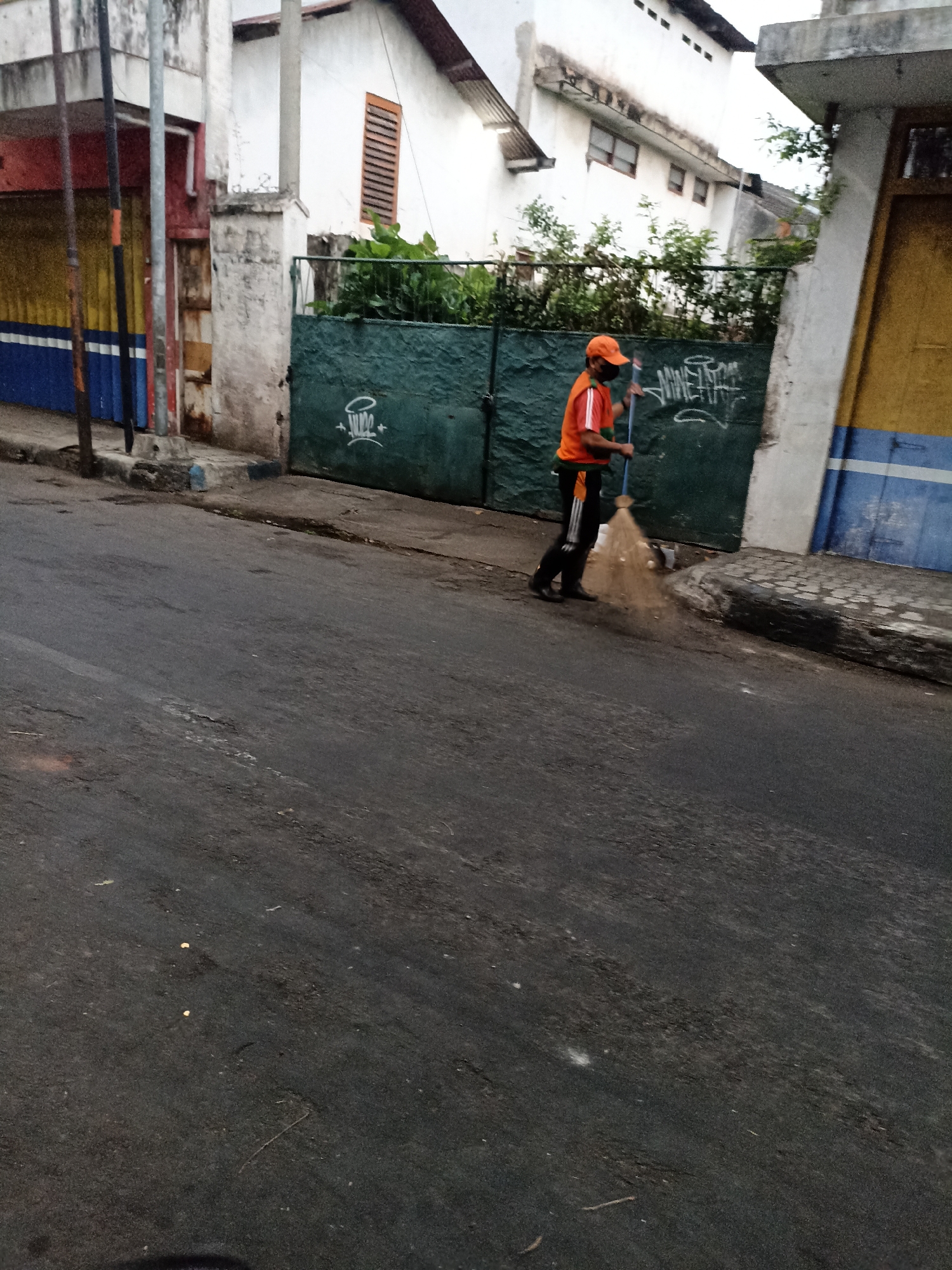 An individual wearing an orange shirt, black pants with white stripes, and a black face mask is sweeping the asphalt street with a broom. The person is standing near a dark green metal gate with graffiti on it, which is adjacent to a building with a white facade and a dark roof. The street appears to be on an incline, and there is a sidewalk with cobblestone paving to the right of the gate. The overall environment suggests an urban setting in Lumajang, Indonesia.