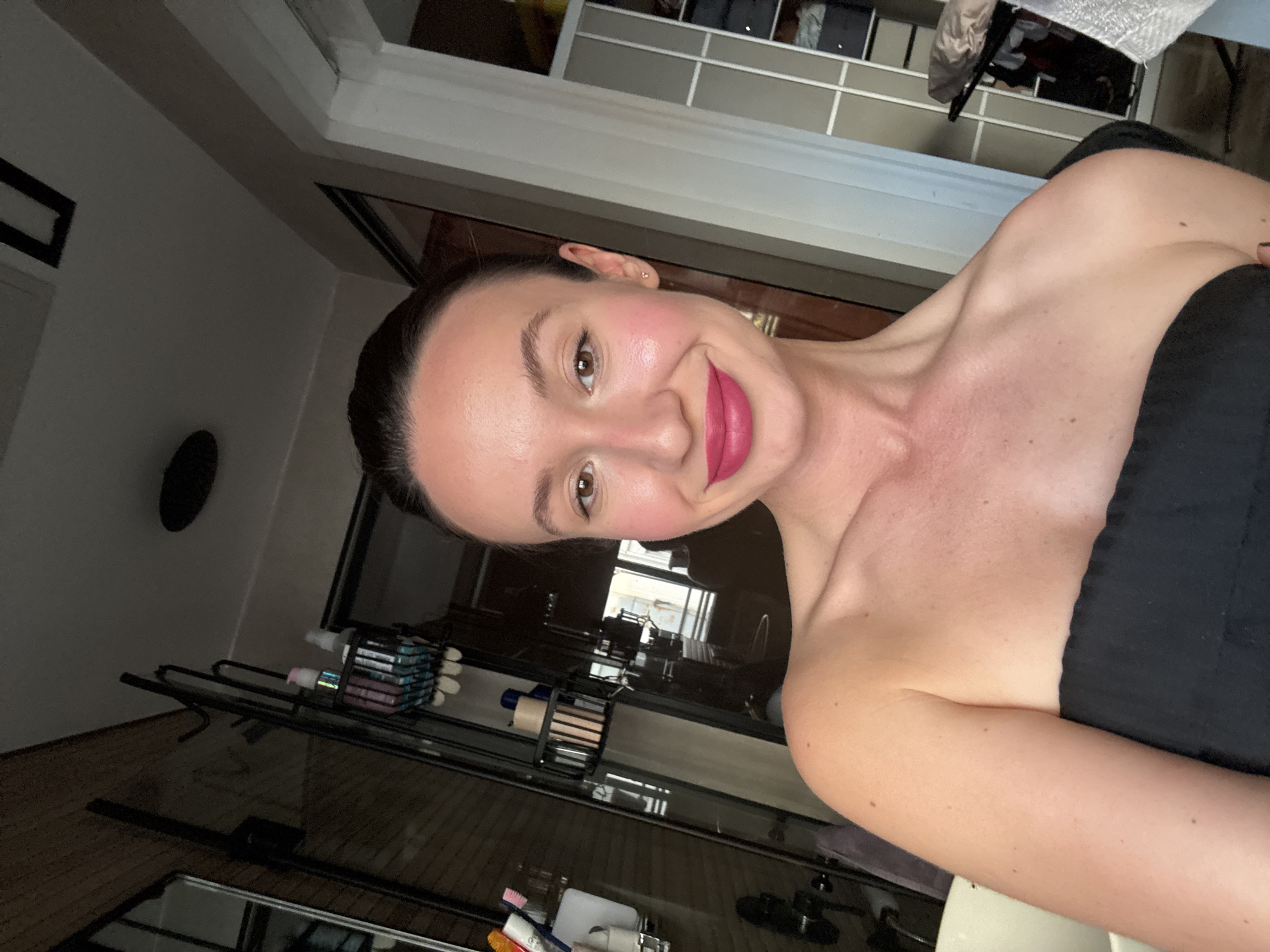 A woman poses for a selfie indoors. She has fair skin, brown eyes, and dark hair pulled back. She wears a strapless black top and bright pink lipstick. The background includes a partially visible wardrobe or shelving unit and a glass-enclosed shelving unit. The location is presumed to be in Tel Aviv, Israel, though no elements in the photo directly confirm it.